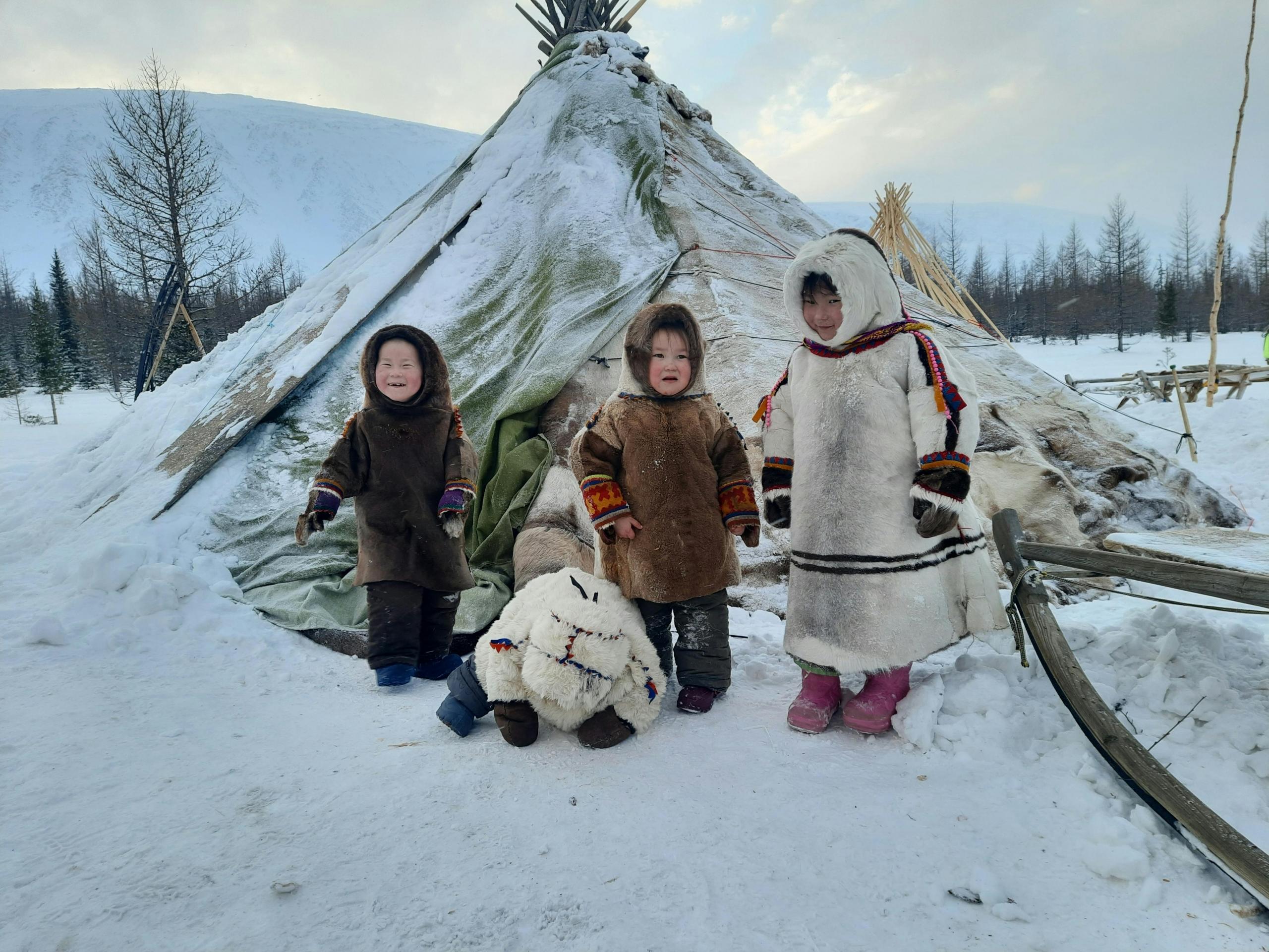 Three children in traditional winter clothes standing in front of a snow-covered tent, with snowy mountains and trees in the background.