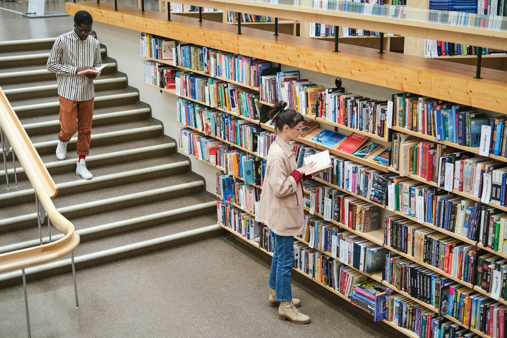 Two people reading books in a library; one stands by shelves, the other walks down stairs holding a book.