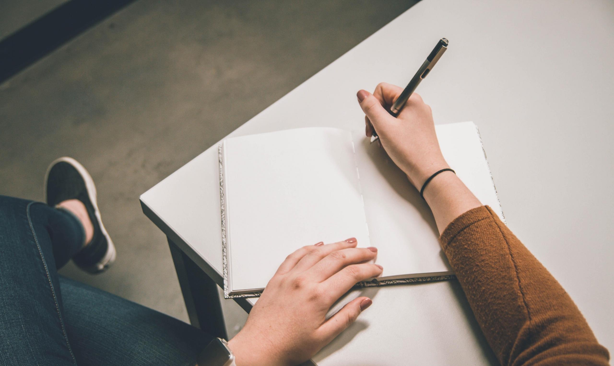 Photograph showing a woman's hands writing on a notebook with white sheets on a desk.