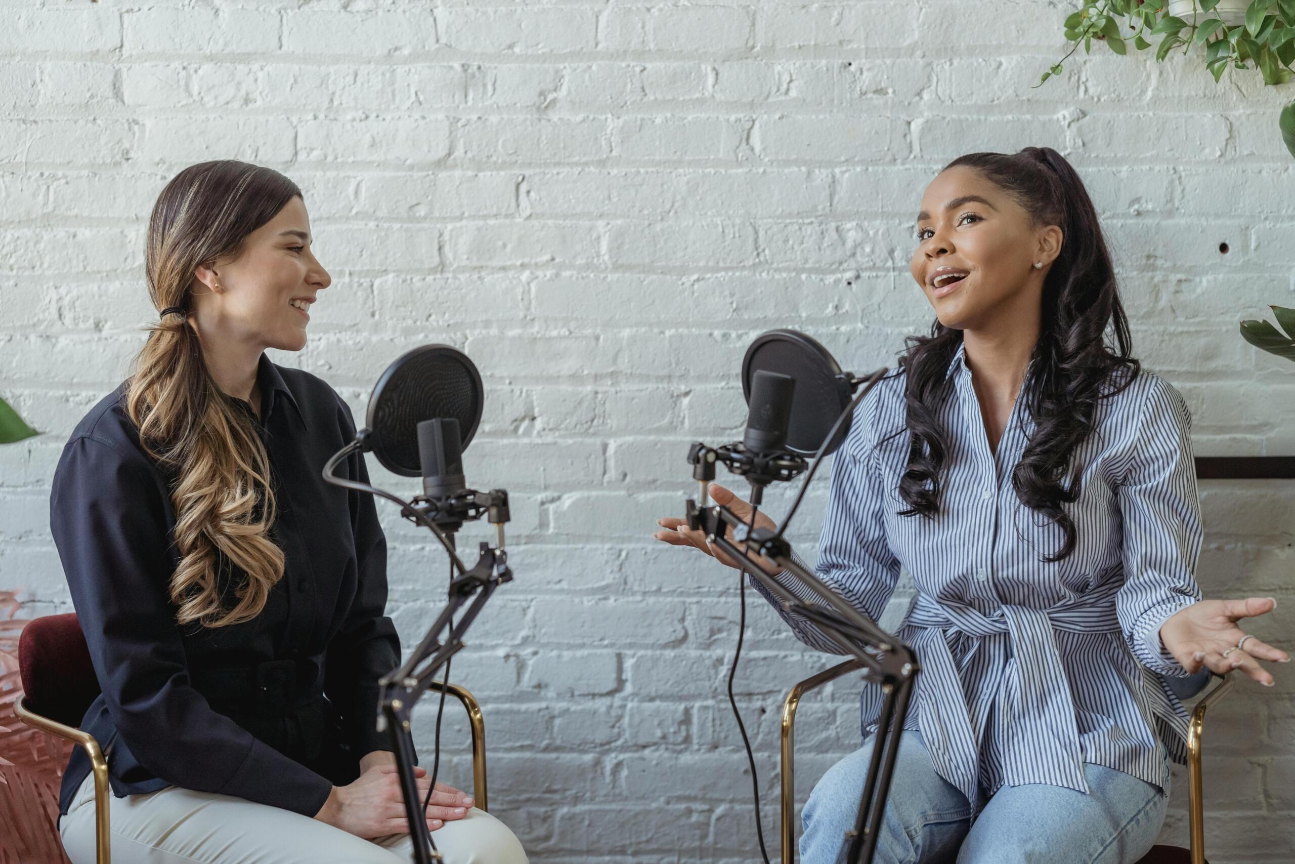 Two women sit in a cozy studio, engaged in conversation, each with a microphone, against a backdrop of a white brick wall and greenery.