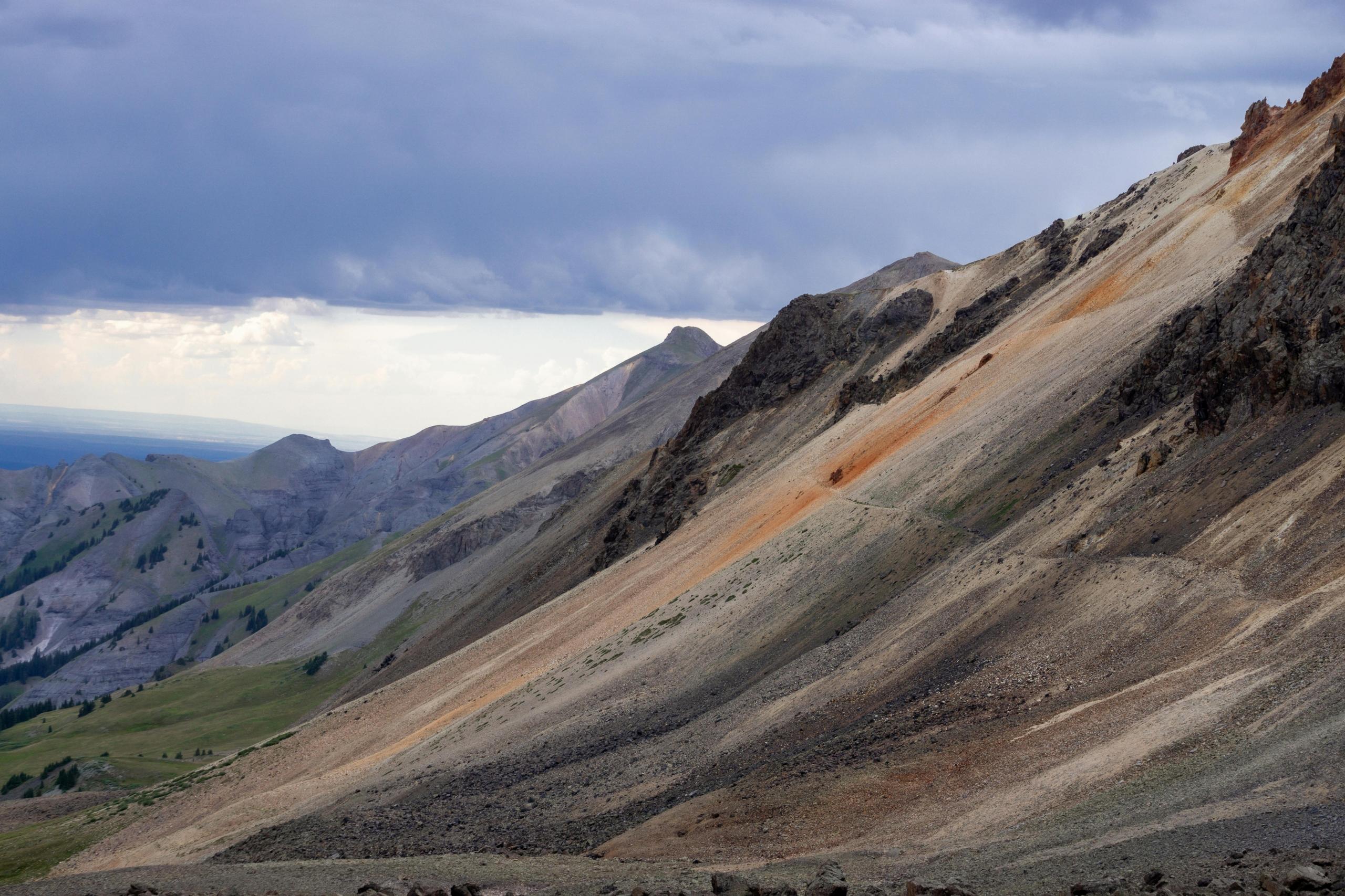 A steep, rocky mountain slope under a cloudy sky, with a mix of green and orange hues marking the rugged terrain.