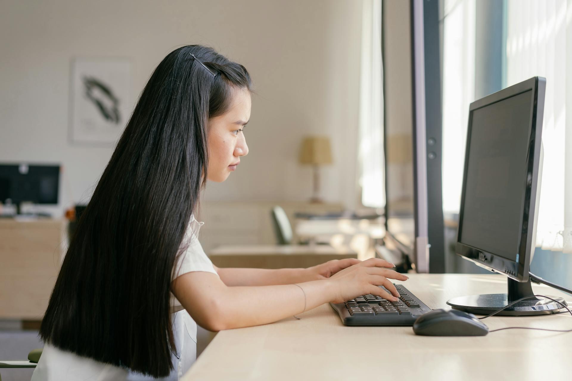 A person with long black hair typing on a keyboard at a desk with a computer monitor in a bright room.