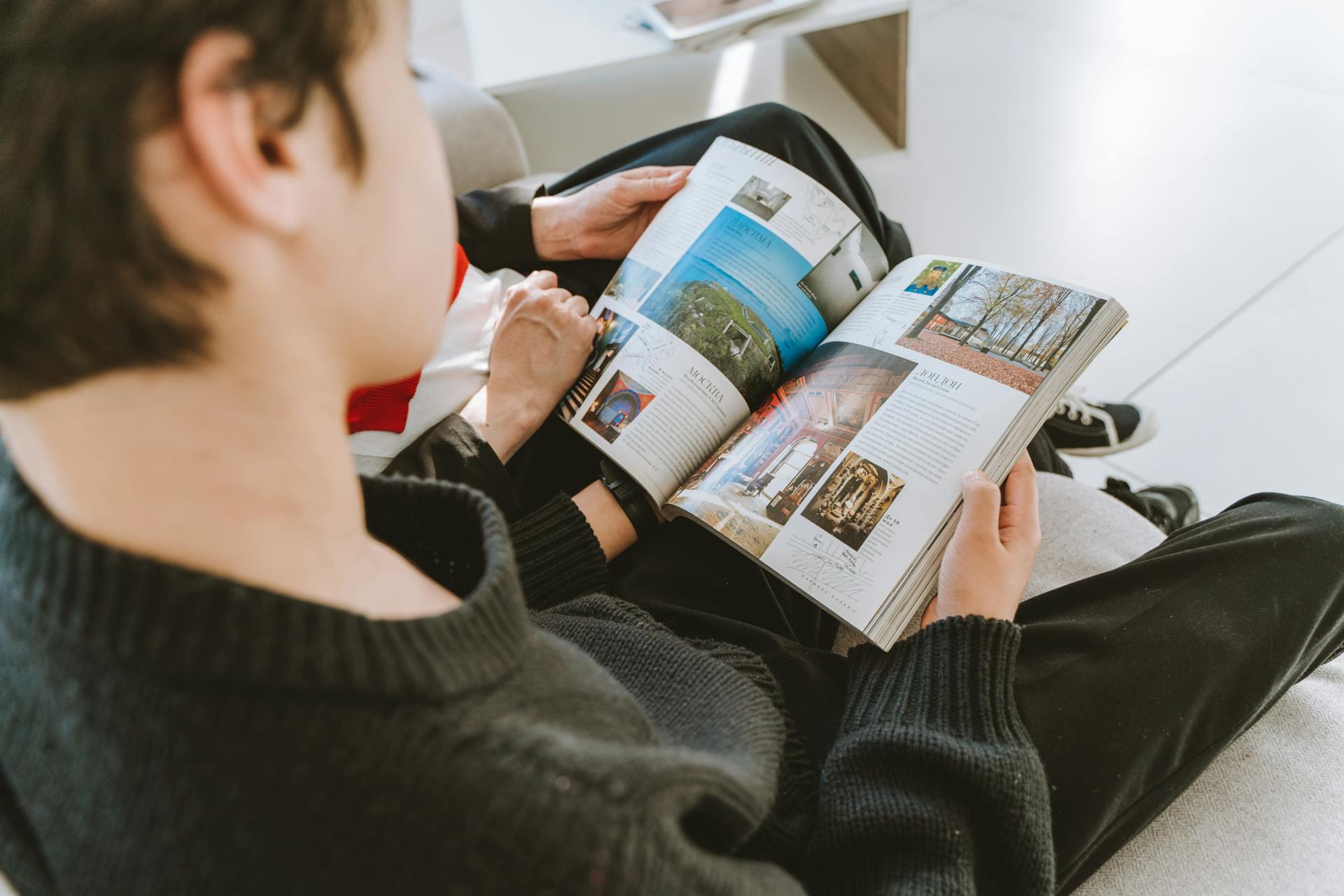 Person reading a magazine with images and text, sitting on a couch with another person partially visible.