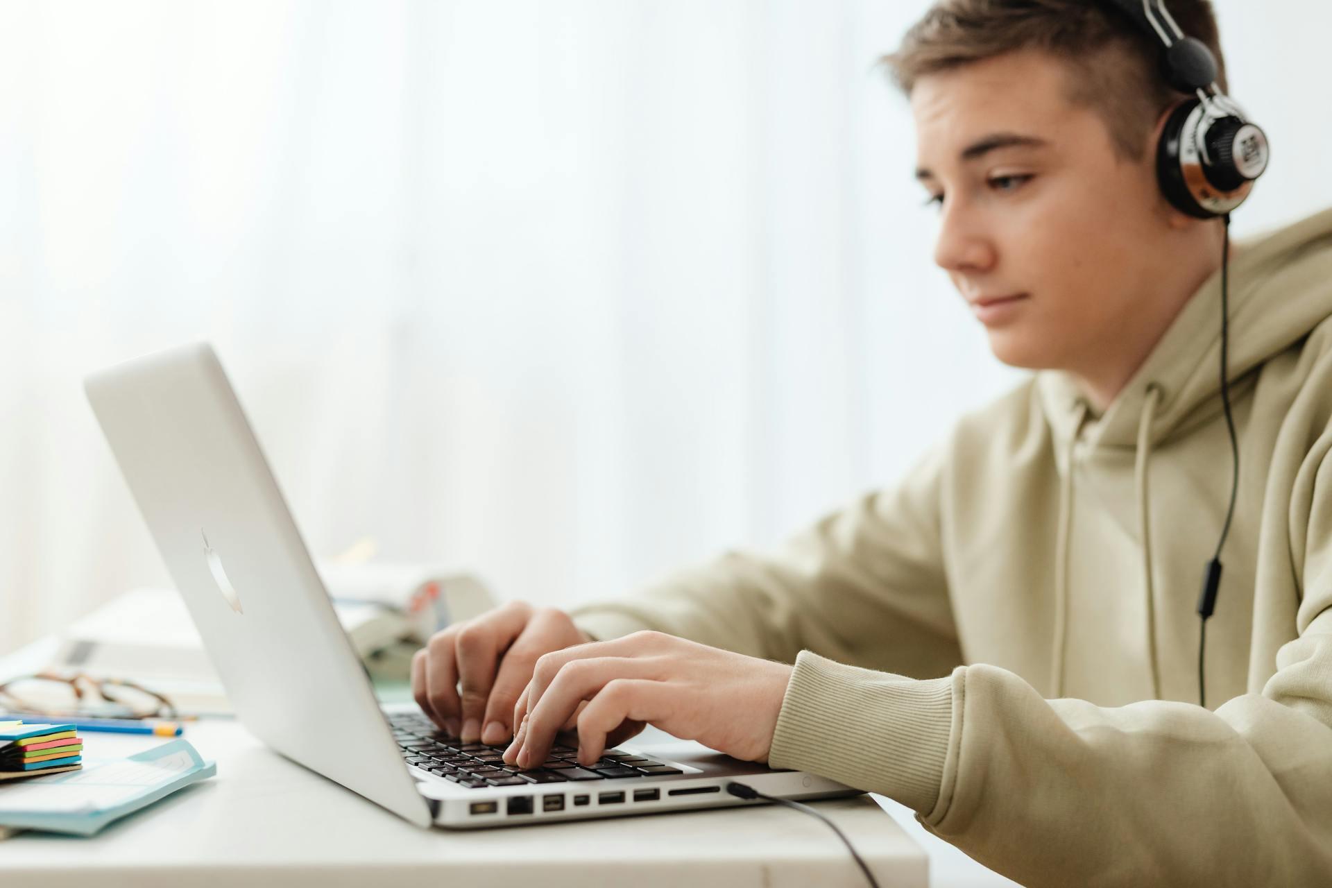 Person wearing headphones and a hoodie typing on a laptop at a desk with papers and stationery nearby.