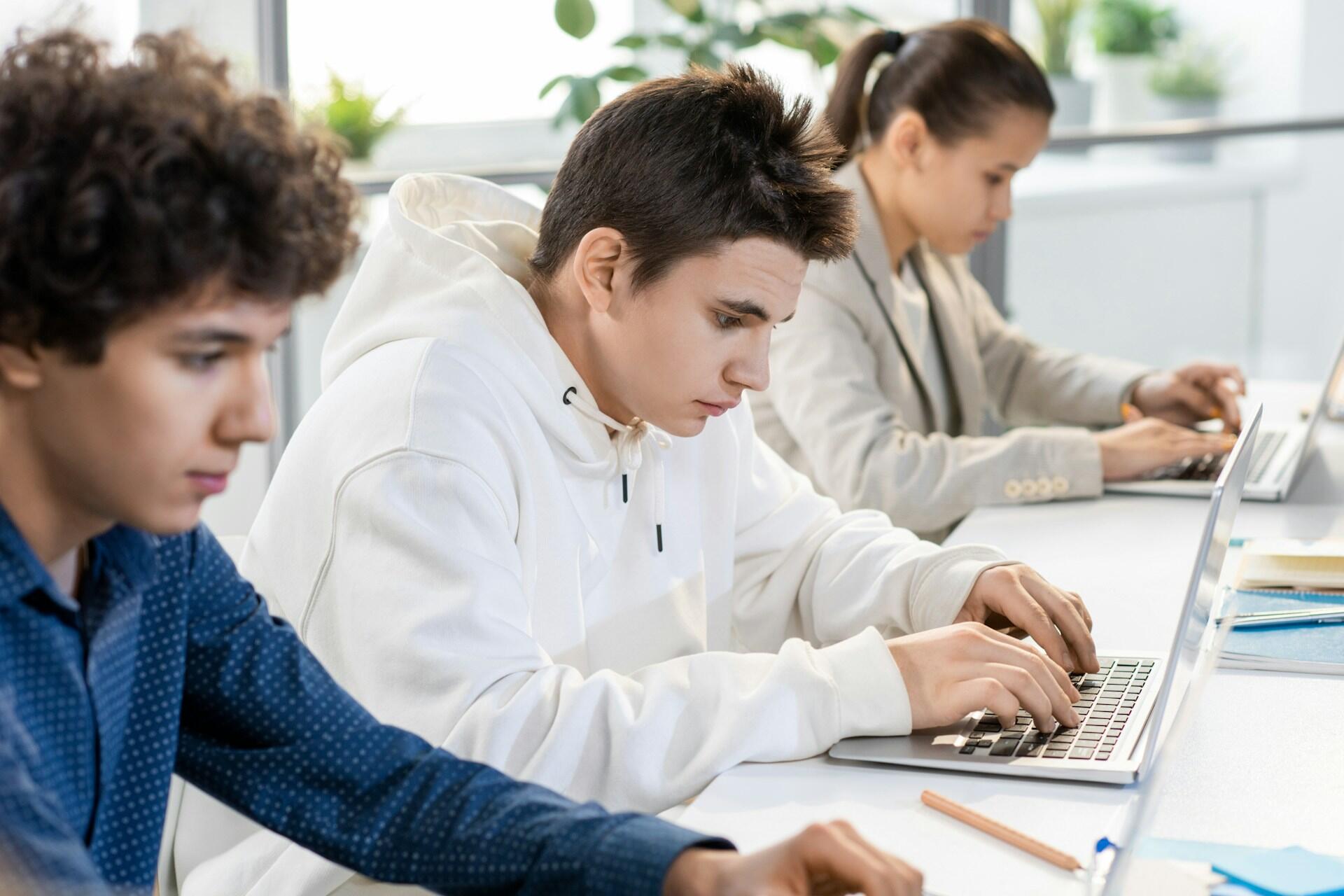 Three young people sitting in a row, focused on typing on laptops at a bright, modern workspace.