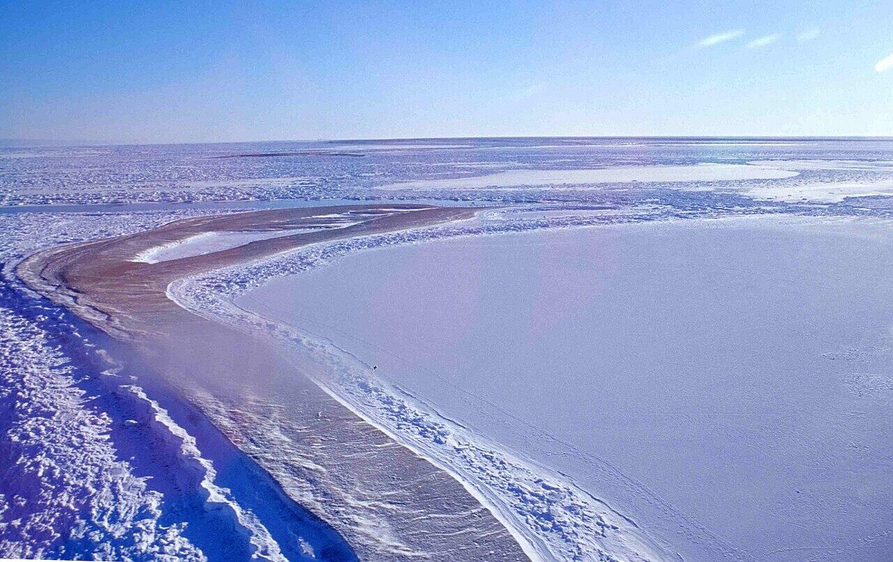 Frozen icy landscape with snow-covered ground under a clear blue sky, featuring frozen water bodies and icy ridges.