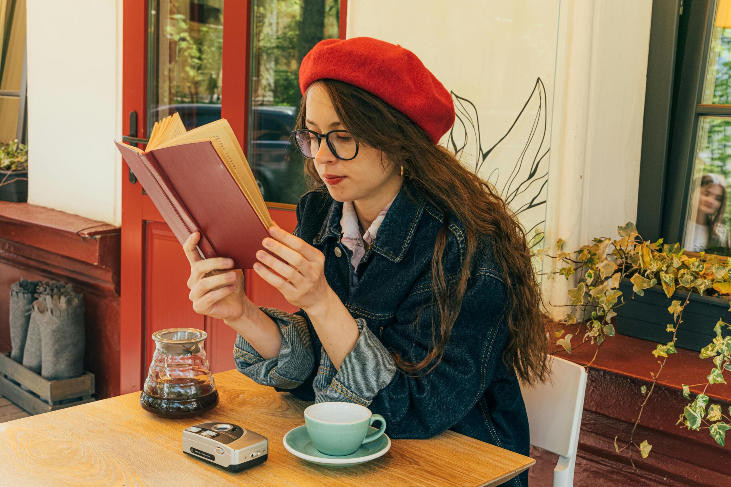A person in a red beret sits at a table in a café, reading a book, with coffee and a vintage camera nearby.