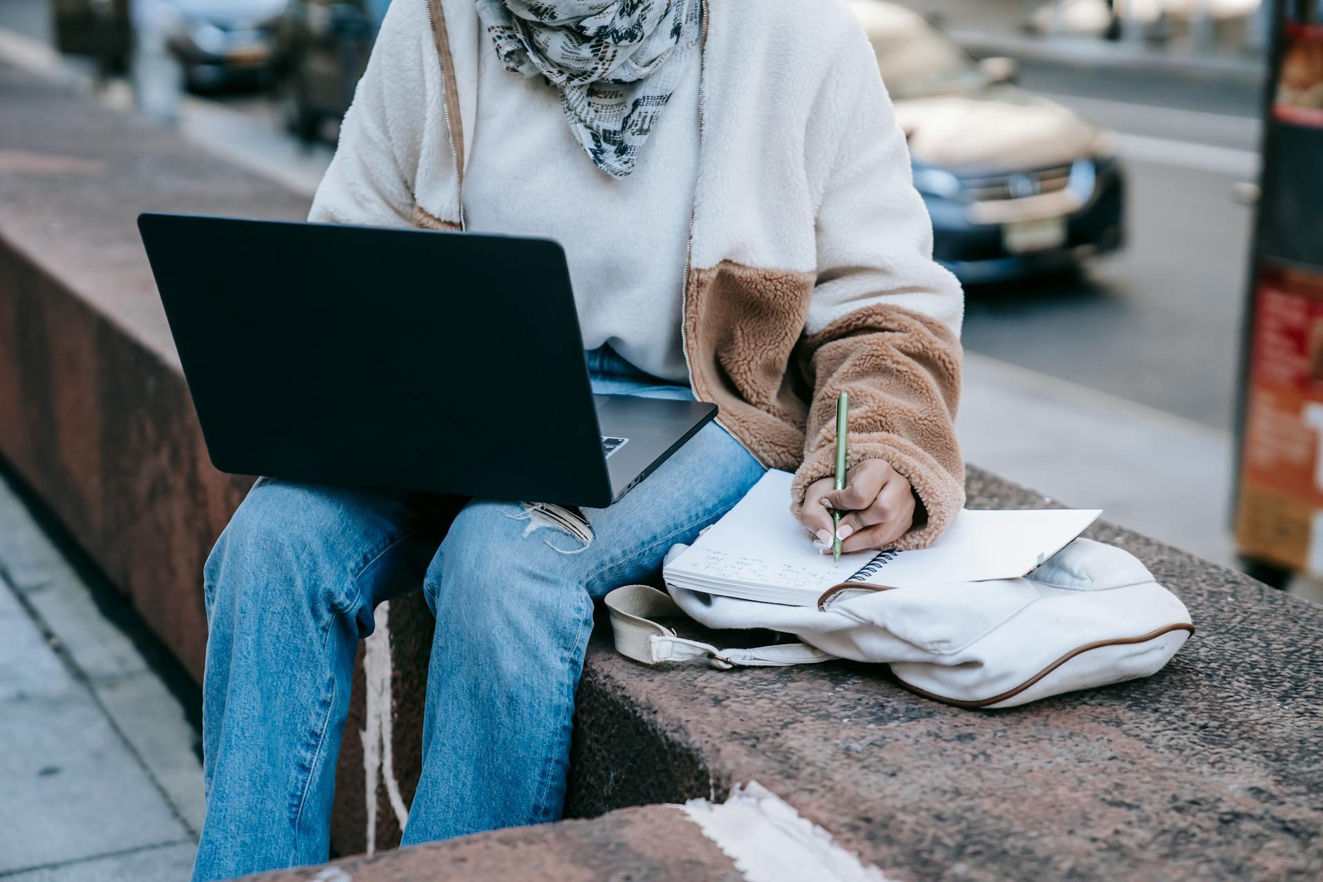 Person sitting on a bench outdoors, using a laptop on their lap and writing in a notebook placed on a bag beside them.