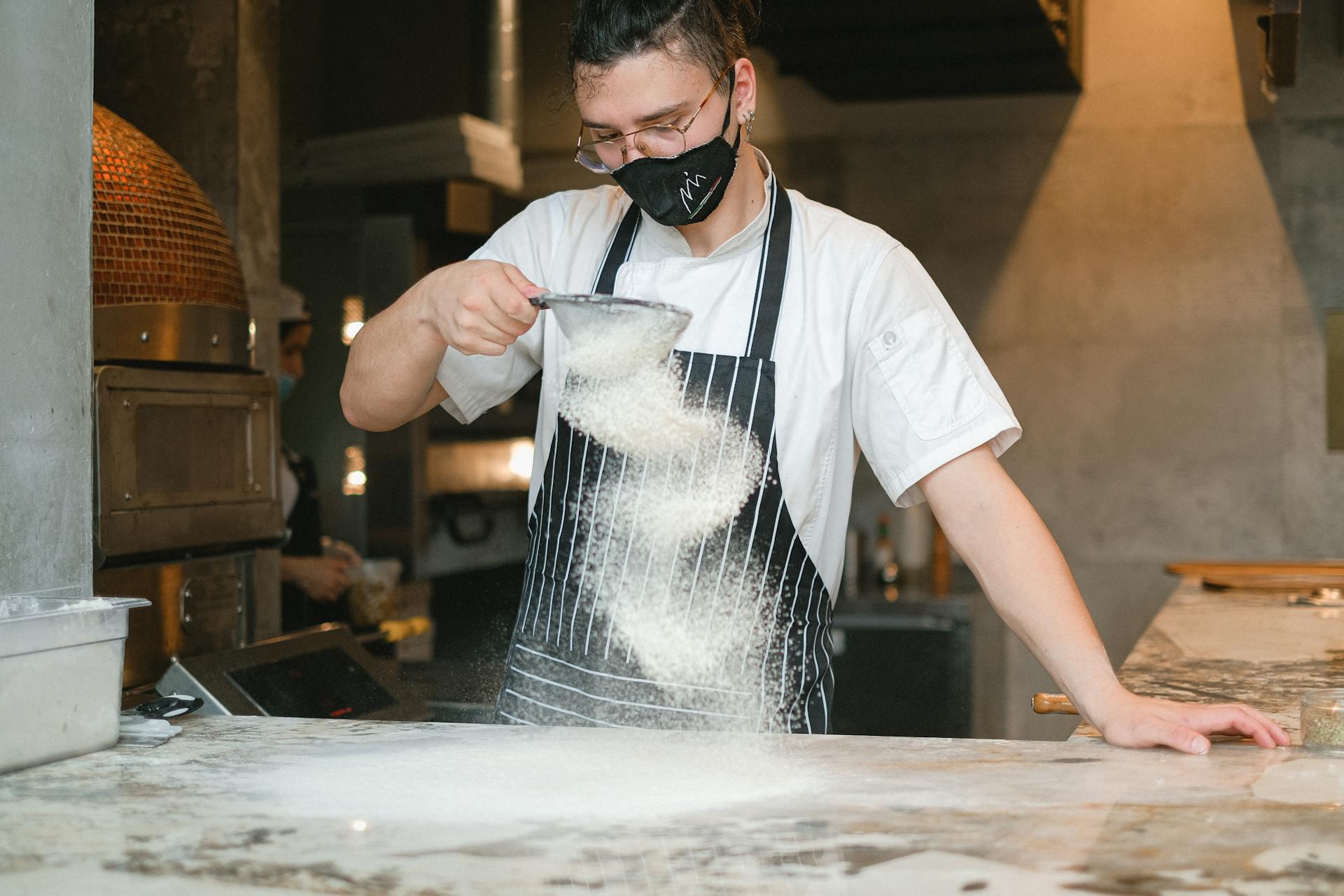Photograph showing a young uniformed pastry chef wearing a mouth cover, pouring flour on a smooth surface. In the background we can see a kitchen.