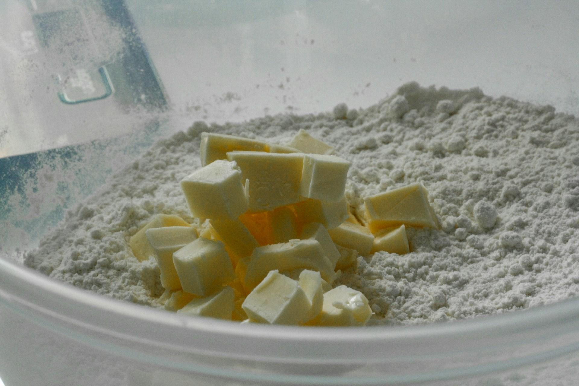 The photograph shows flour sifted with a cube of butter in a glass bowl.