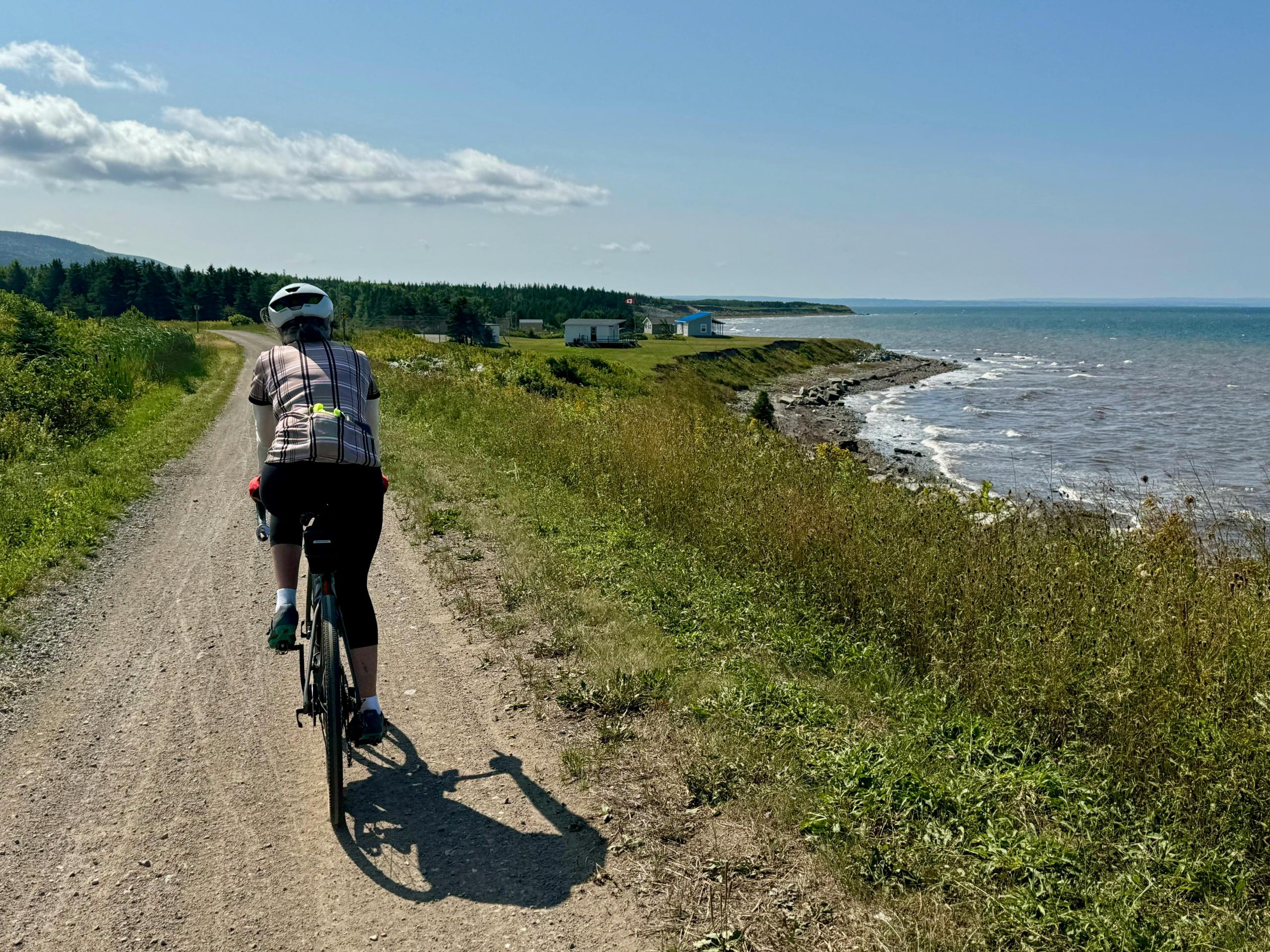 a man cycles along a trail by the coast. The sky is blue and it is sunny.