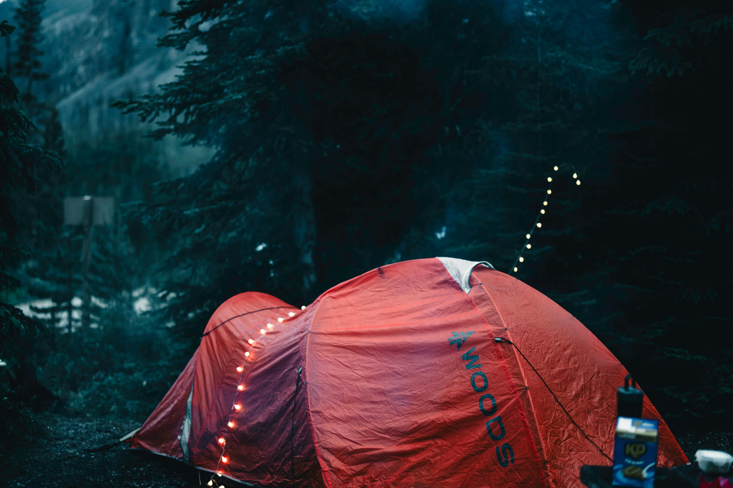 A red tent illuminated by string lights set up in a forested campsite in Banff National Park during dusk.