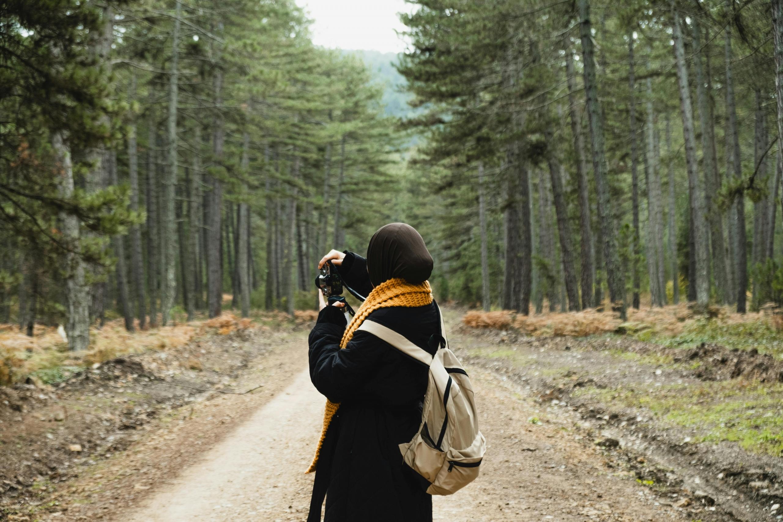 a woman in a coat and scarf takes photos of a forest.