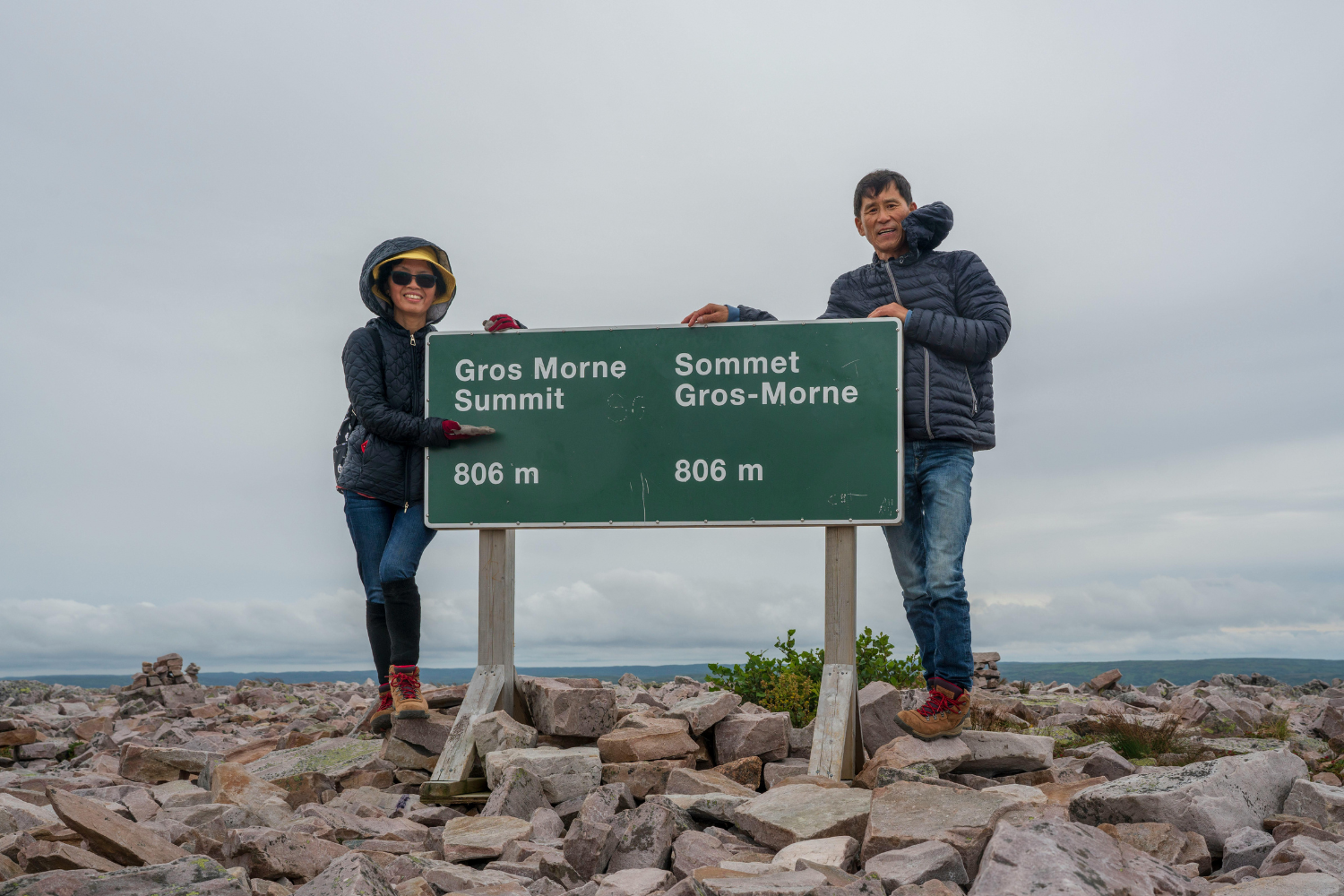 Two people standing beside the summit sign of Gros Morne, reading “Gros Morne Summit 806 m.