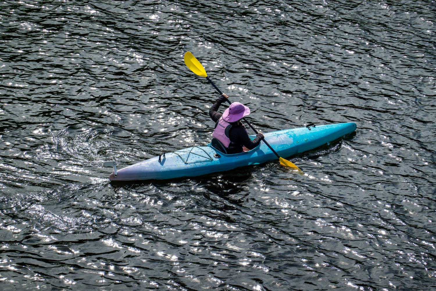 A person kayaking on calm, reflective waters, wearing a purple hat and life jacket, surrounded by rippling waves.