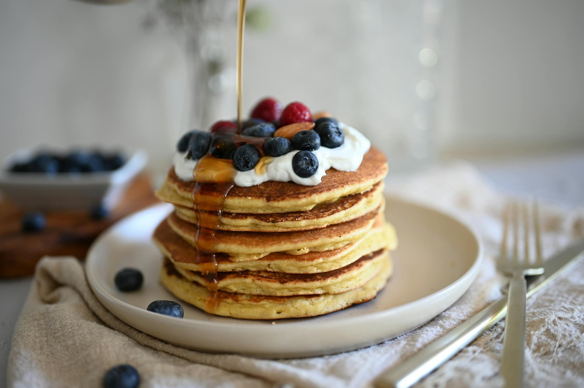 Photograph showing pancakes stacked on a white plate with maple syrup and berries.