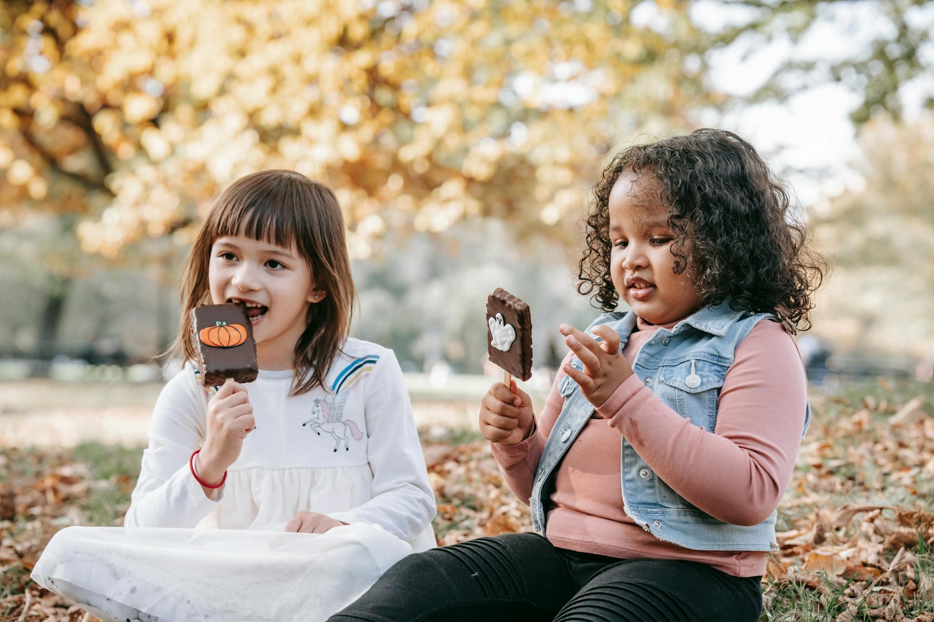 The picture shows two girls enjoying Halloween chocolate bars in a park with the Fall leave-fall in the background. 