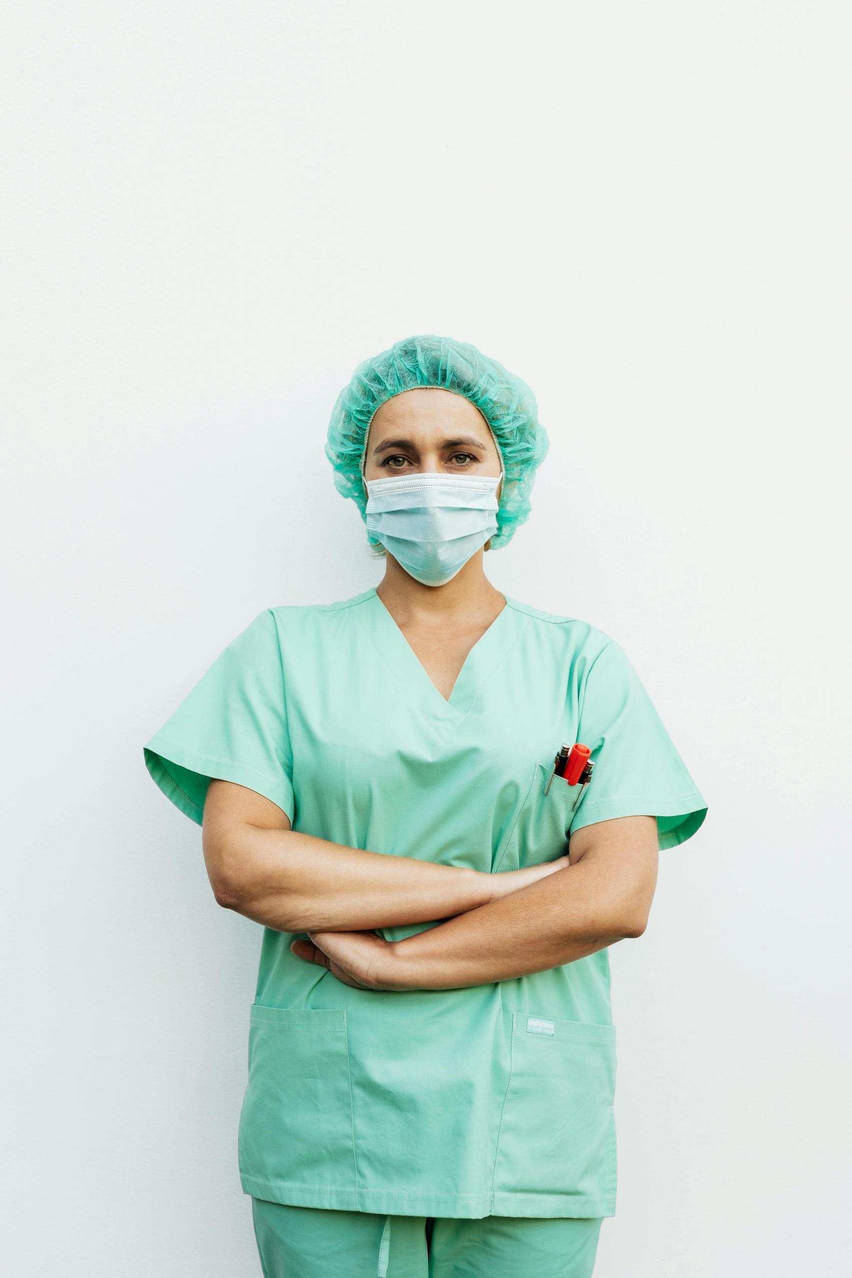 a nurse wears a mask and green scrubs. She looks directly at the camera with her arms crossed.