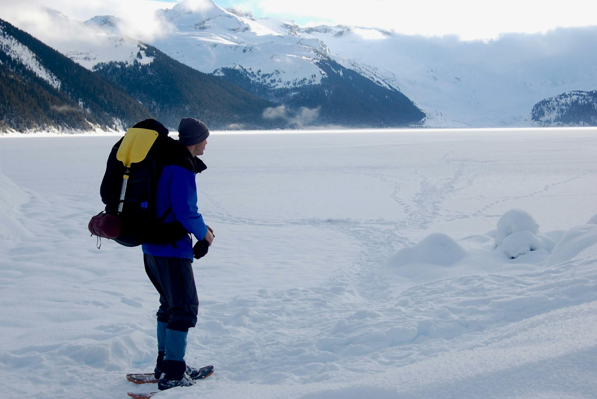 a man carrying a backpack while snowshoeing with snowcapped mountains in the background.