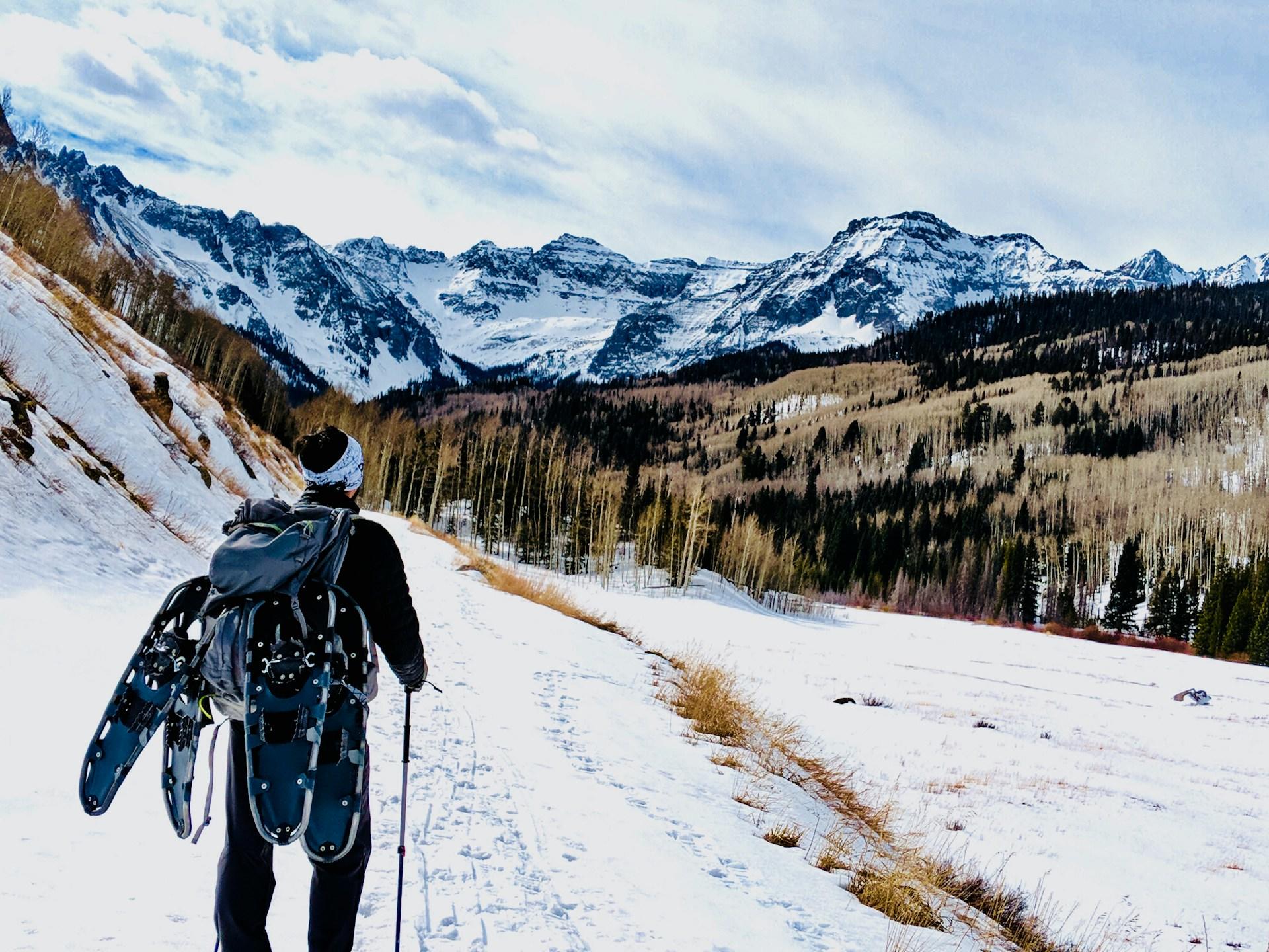 Person with a backpack and snowshoes hiking towards snowy mountains.