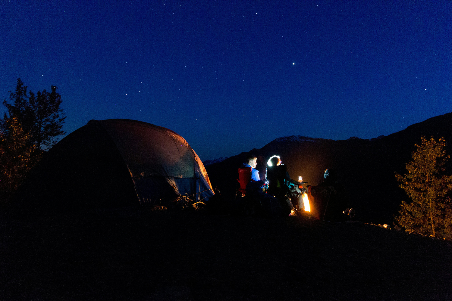 A camping scene at night with a glowing tent, campers around a fire, and a sky full of stars in Jasper National Park.
