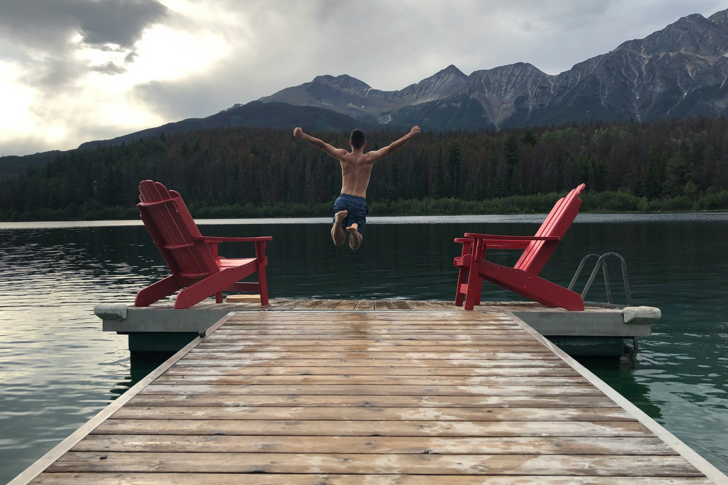 A dock with two iconic red Adirondack chairs and a person jumping into a clear lake, framed by forested mountains and cloudy skies in Jasper National Park.
