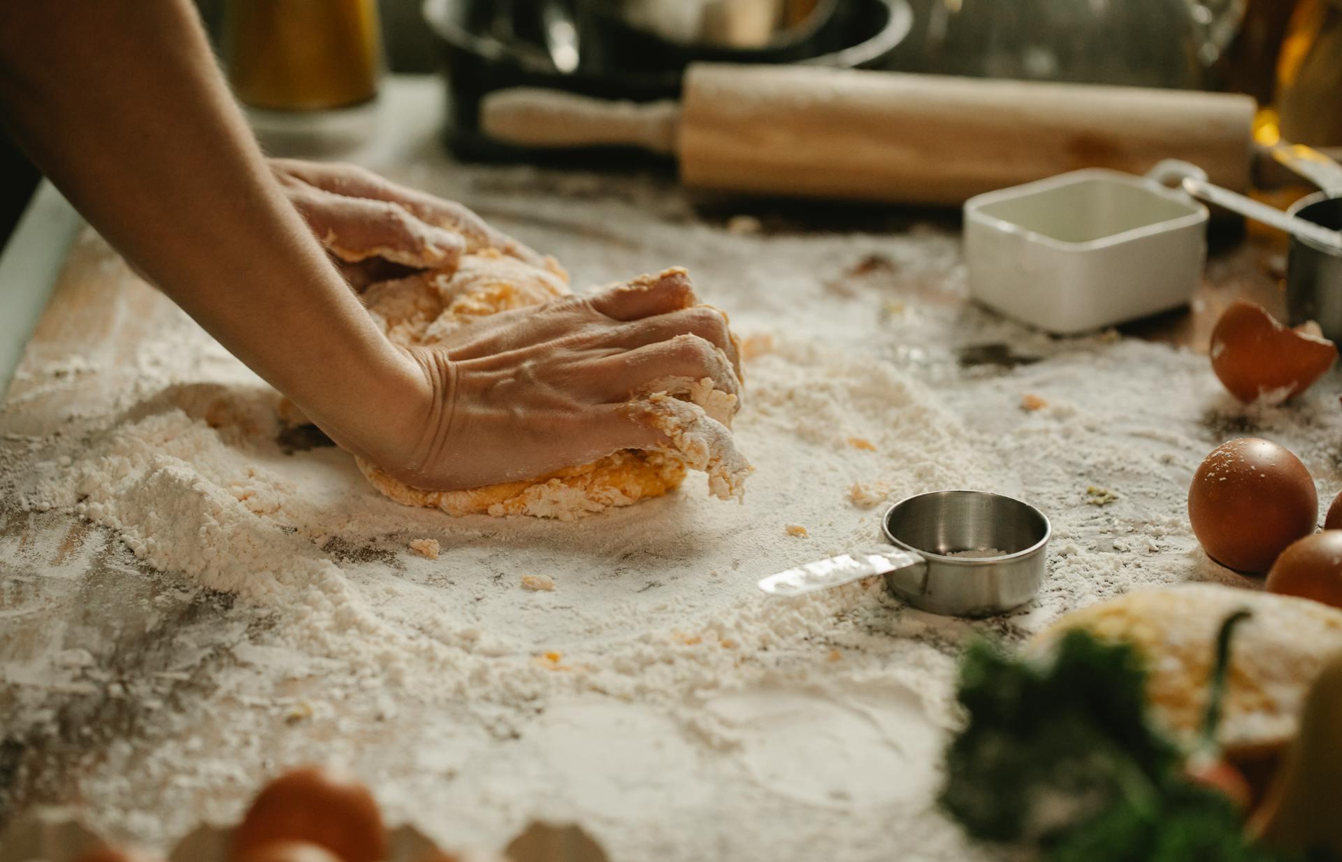 The photograph shows a person kneading dough with his hands on a table covered with flour.