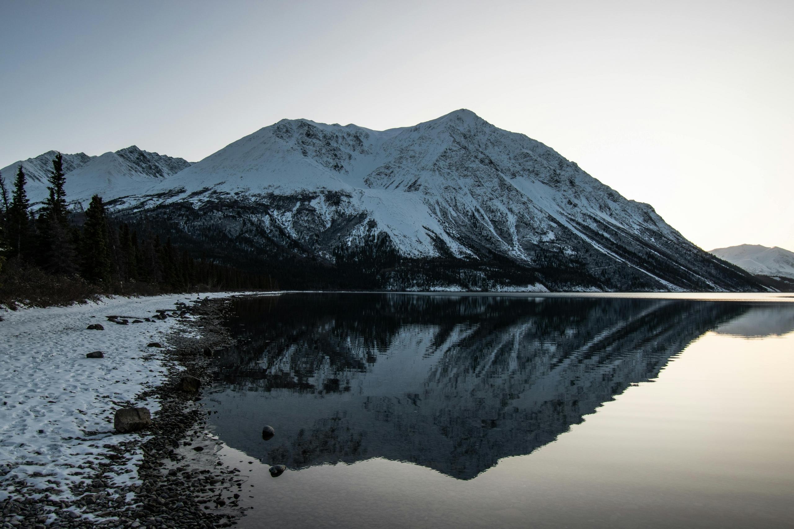 A snowy mountain rises dramatically from a lake. The beach is covered in snow. The mountain is reflecting into the lake.