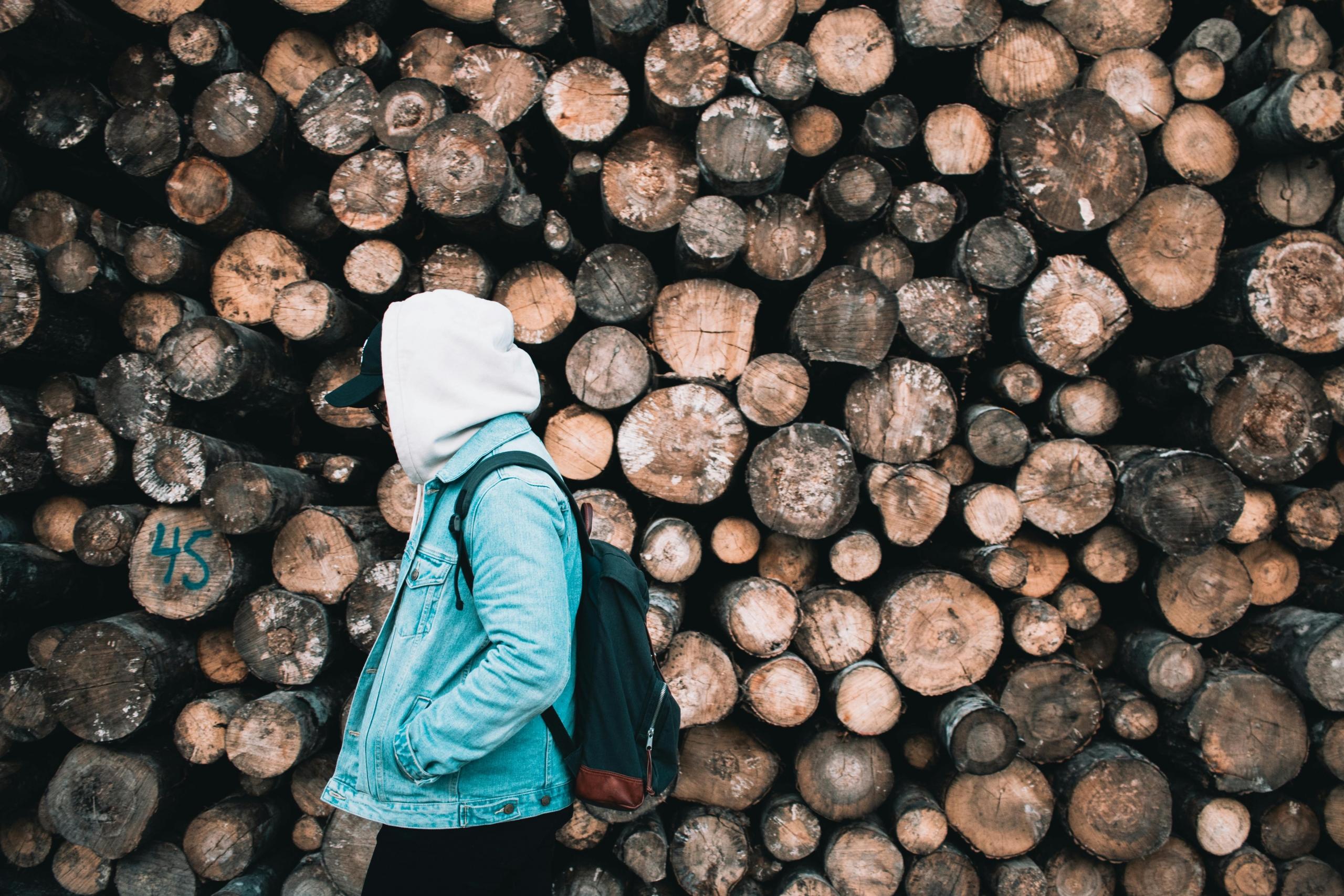 a person in a hoodie and denim jacket walks past a large pile of wooden logs.