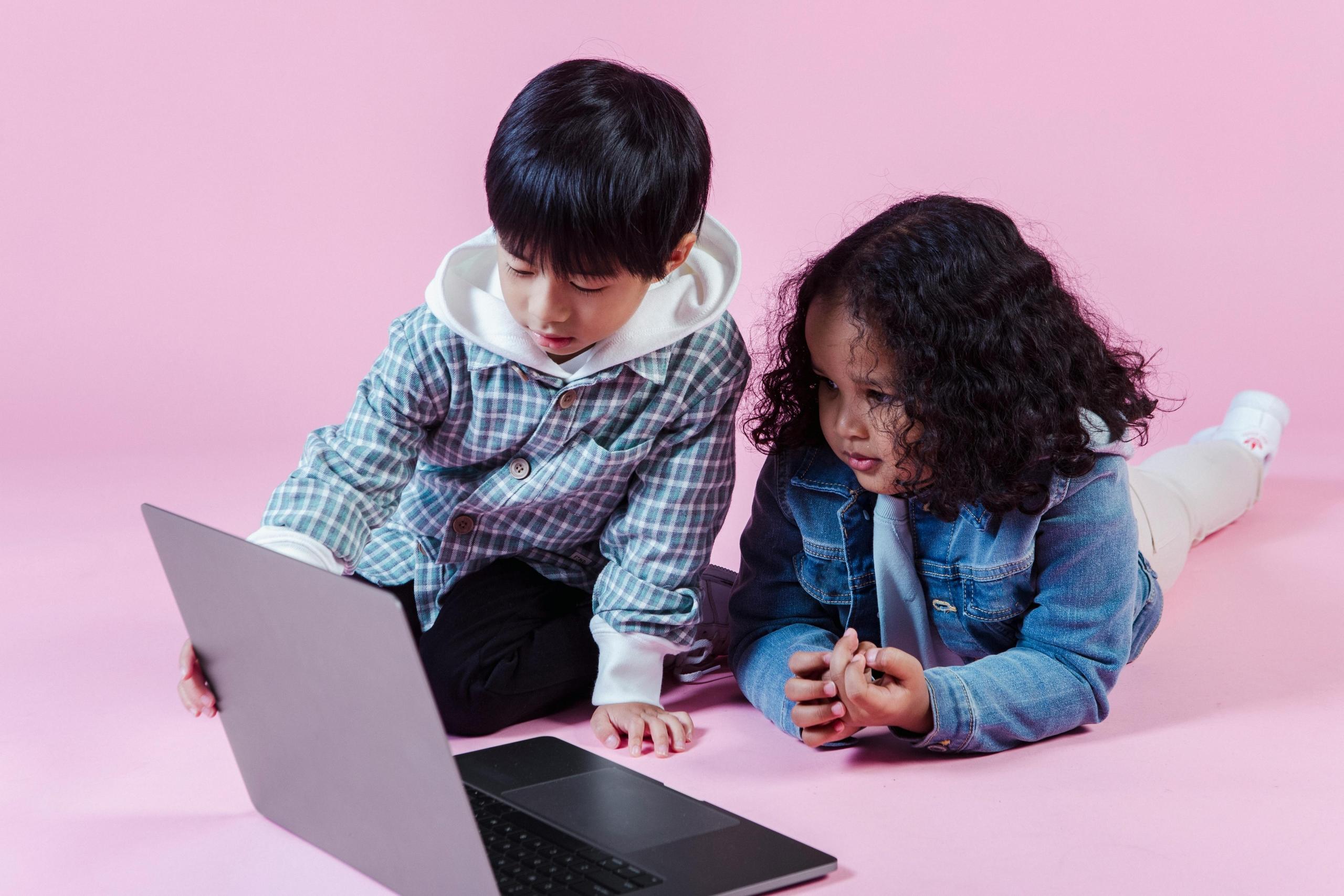 a boy and a girl look at a laptop. The background is pink.