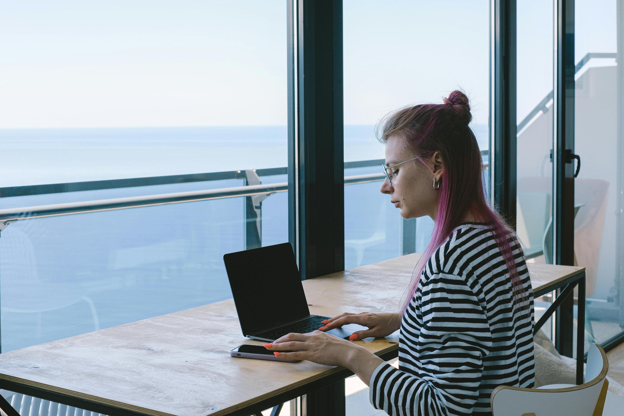 a woman uses a laptop at a desk. The ocean can be seen in the background.