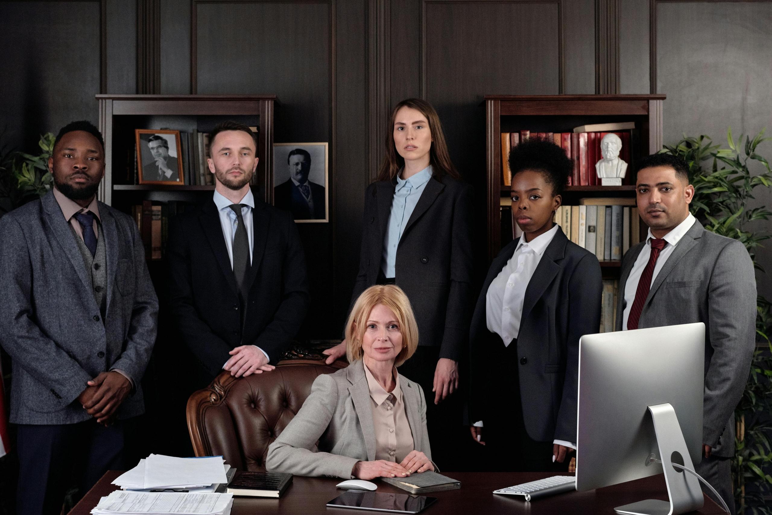 a group of legal professionals from different racial backgrounds look solemnly at the camera and pose around a desk.