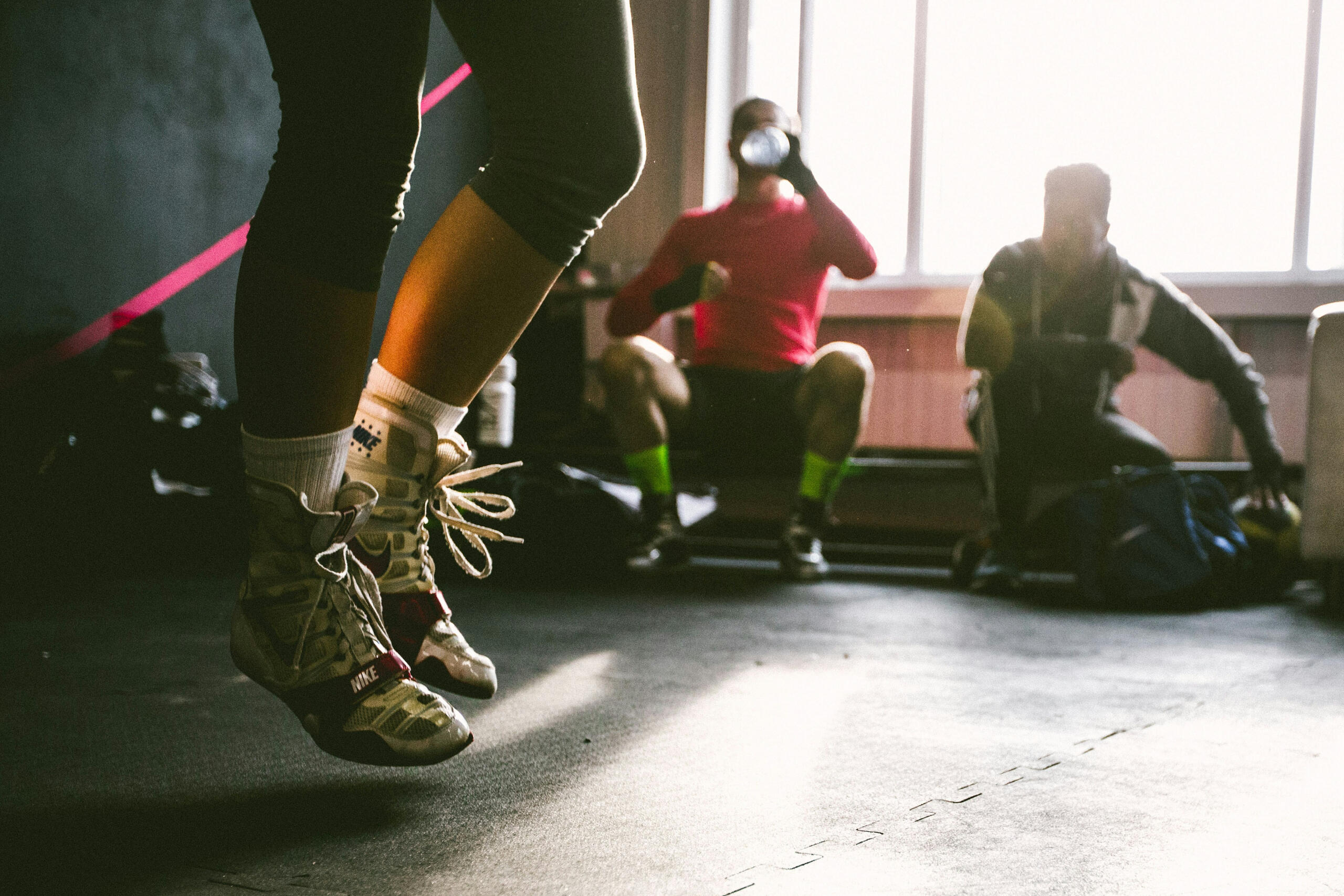 A person jumping rope in a fitness class, with two individuals seated in the background. The room has natural light streaming in through large windows.