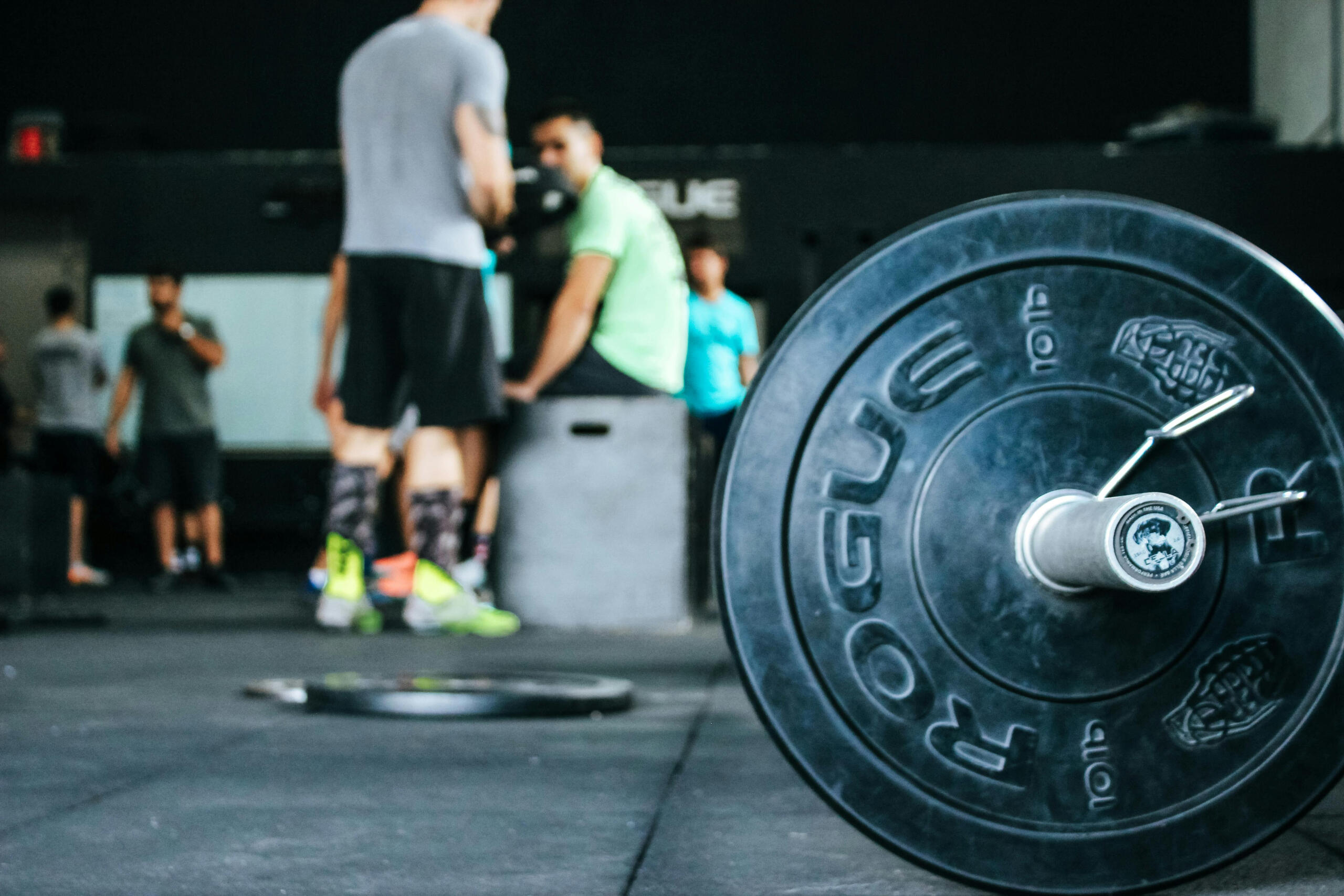 A close-up of a weight plate on the gym floor, with a blurred background of people participating in a fitness class, likely engaging in high-intensity workouts such as CrossFit or strength training.