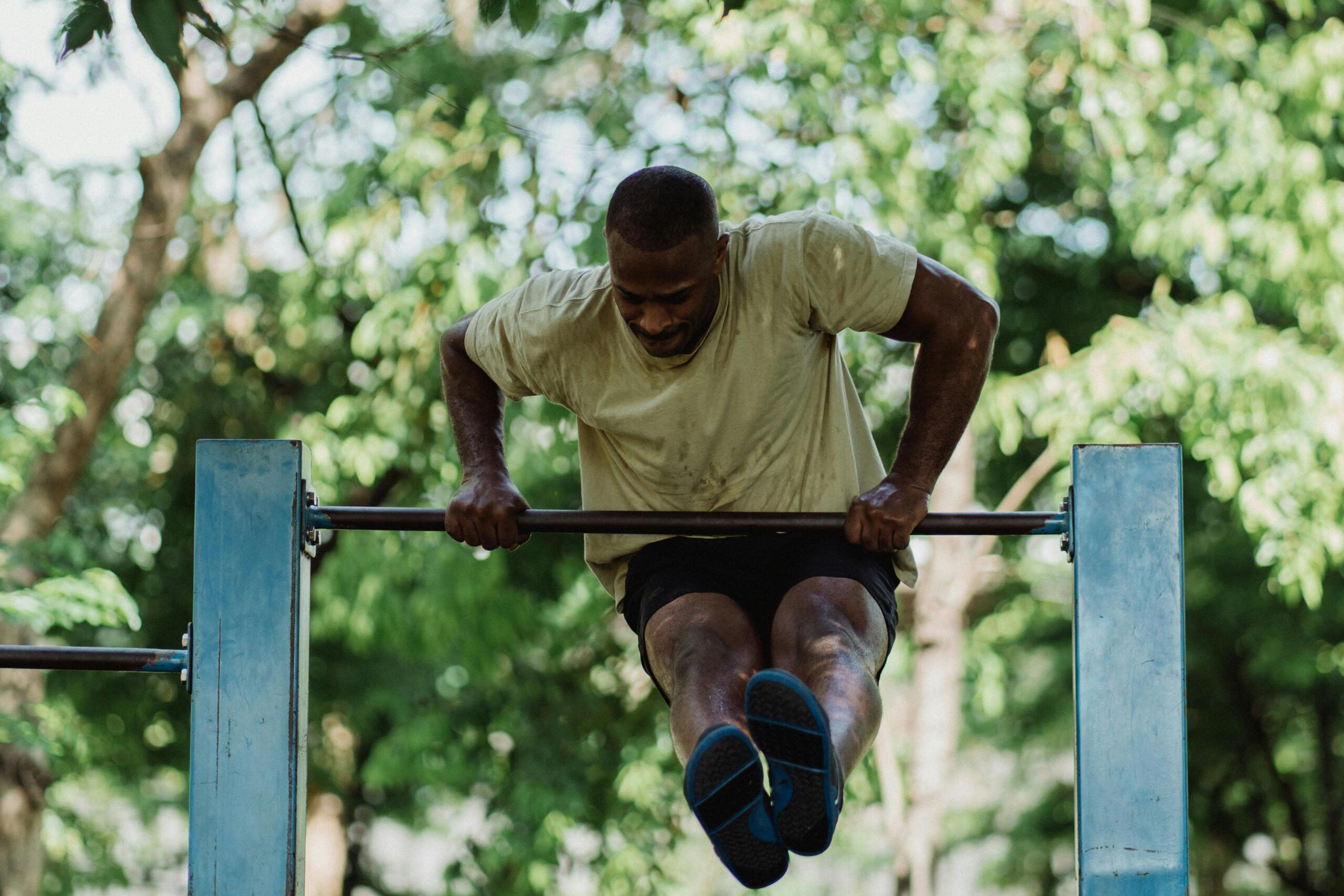 A person in a light-colored t-shirt and shorts is performing a dip exercise on parallel bars outdoors. The background features green trees and natural foliage.