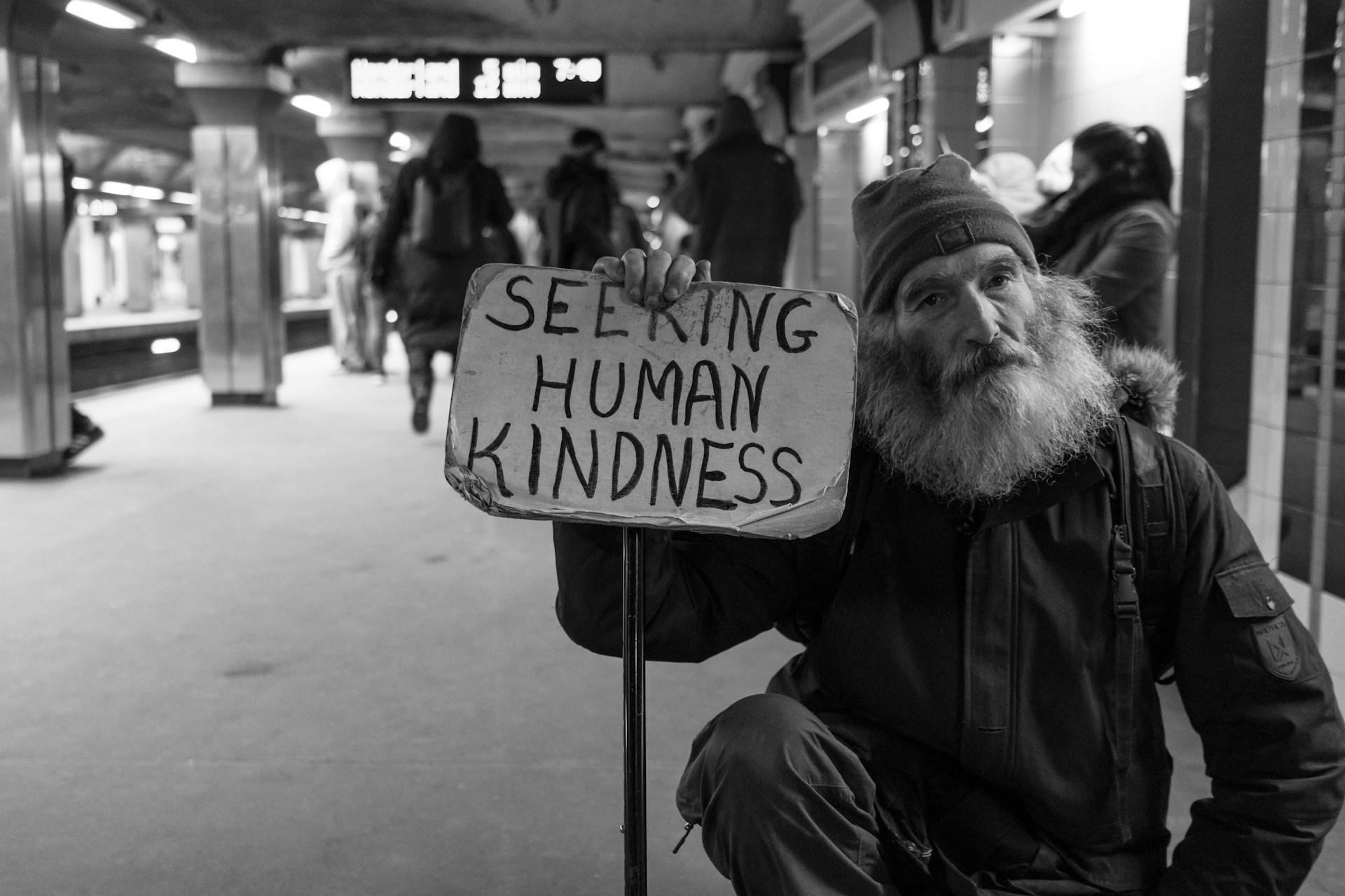 Person holding a sign with "SEEKING HUMAN KINDNESS" in a subway station.