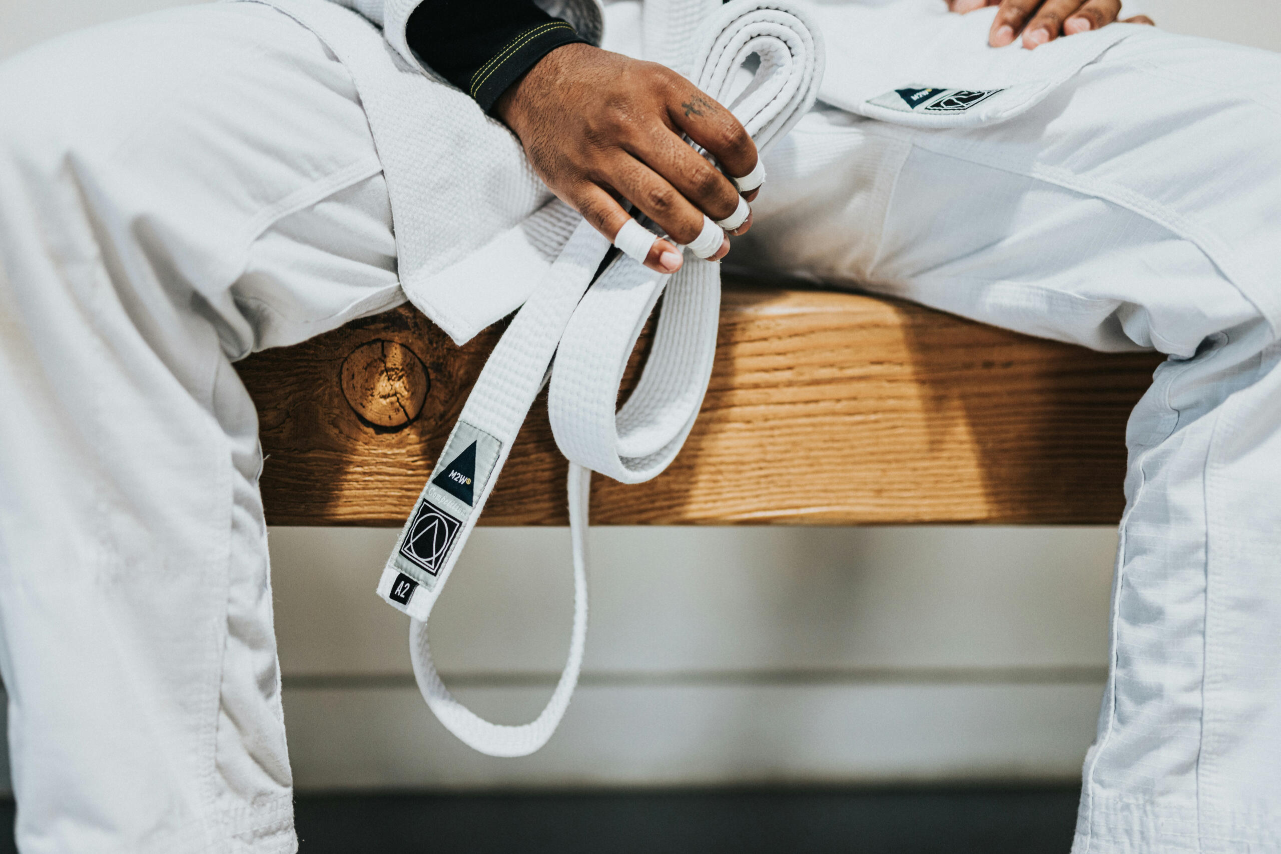 A person holding a Brazilian Jiu-Jitsu gi belt, sitting on a wooden bench, dressed in a traditional martial arts uniform.