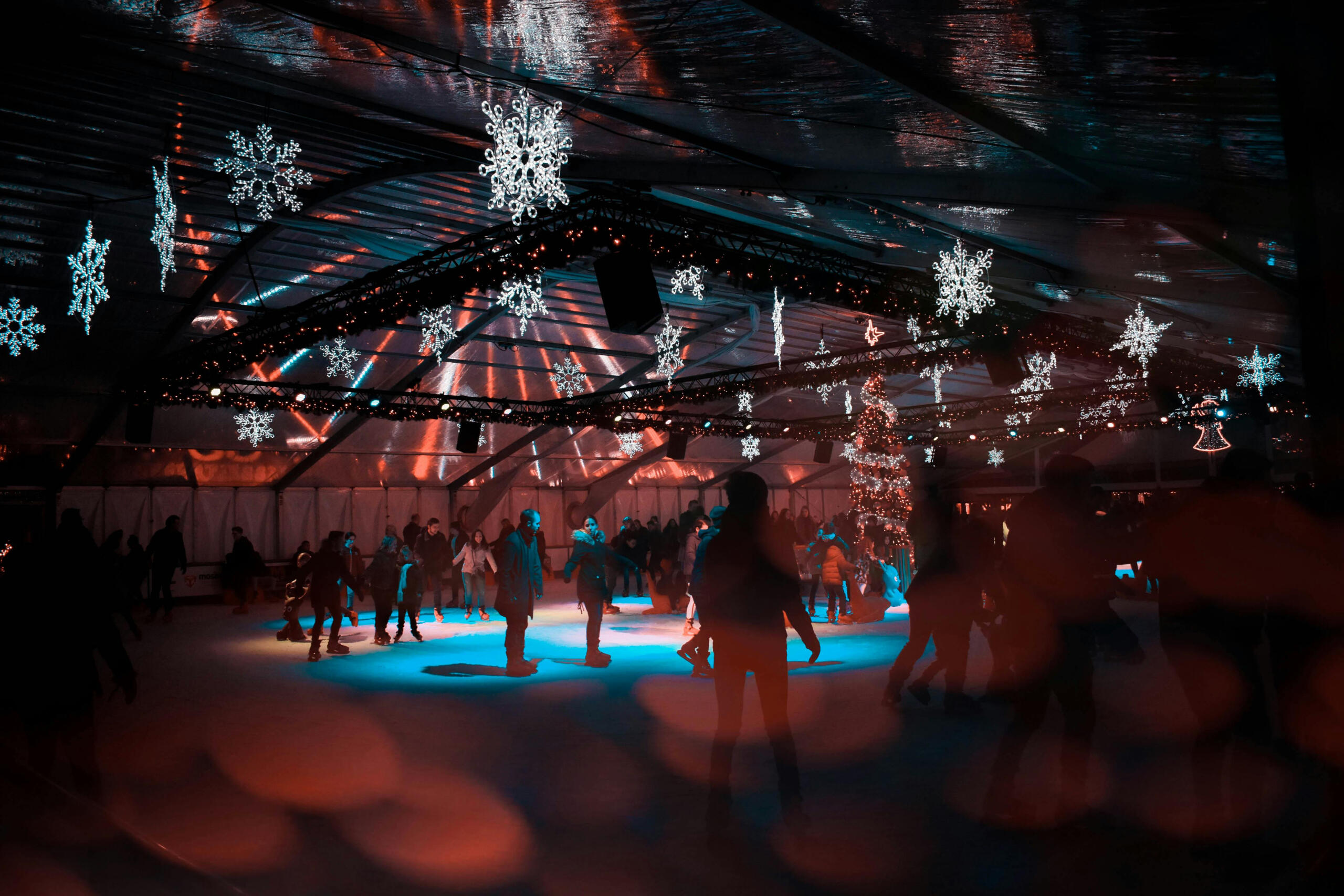 a covered skating rink with illuminated snow flakes and coloured lights. Skaters are on the ice.