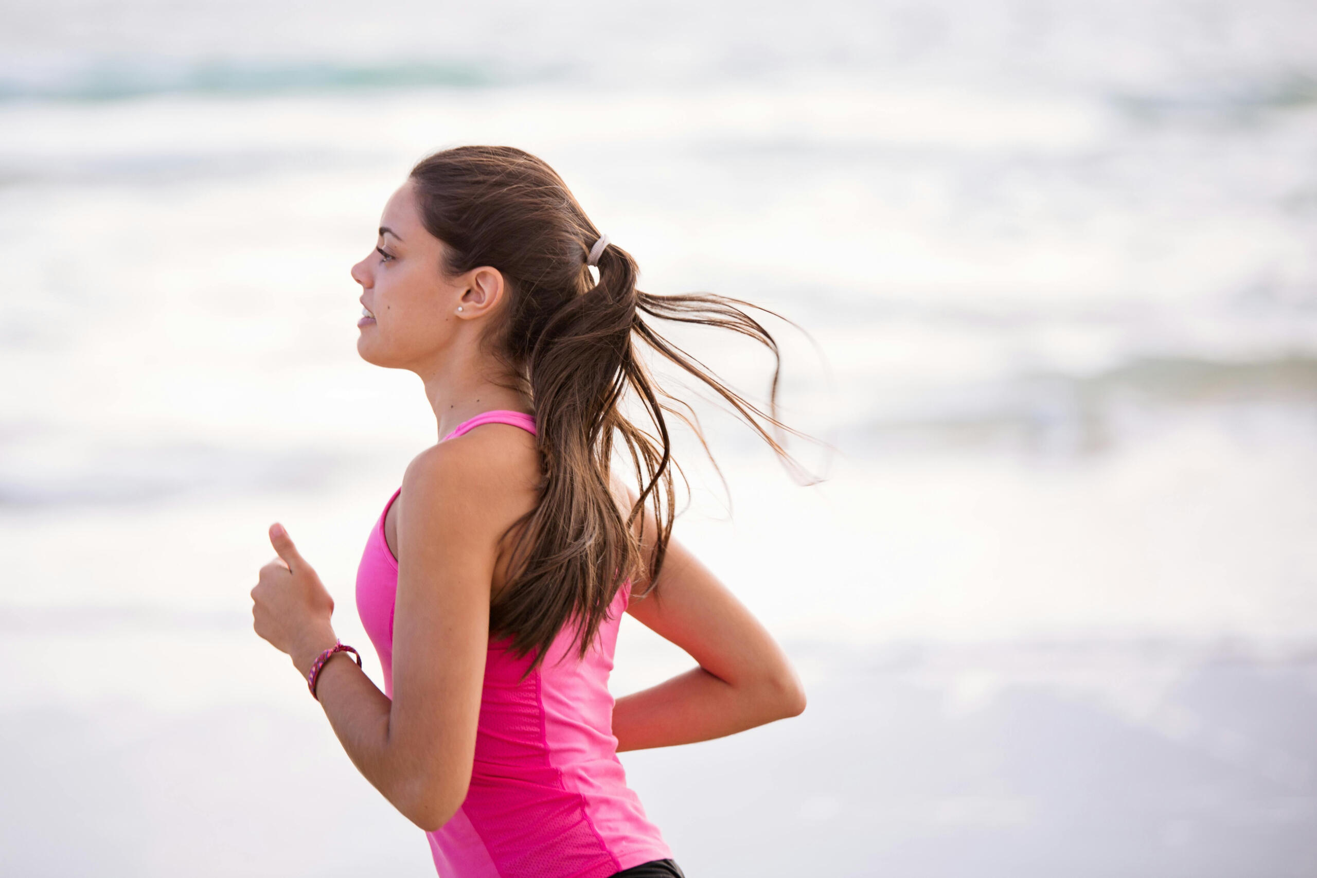 a woman in a pink tank top runs by the ocean. Her hair is in a ponytail.
