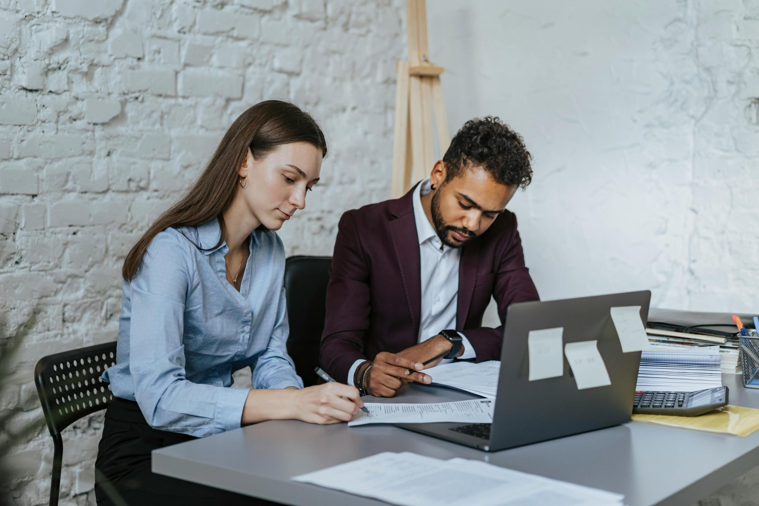 A man and a woman collaborate over a laptop in a professional work environment.