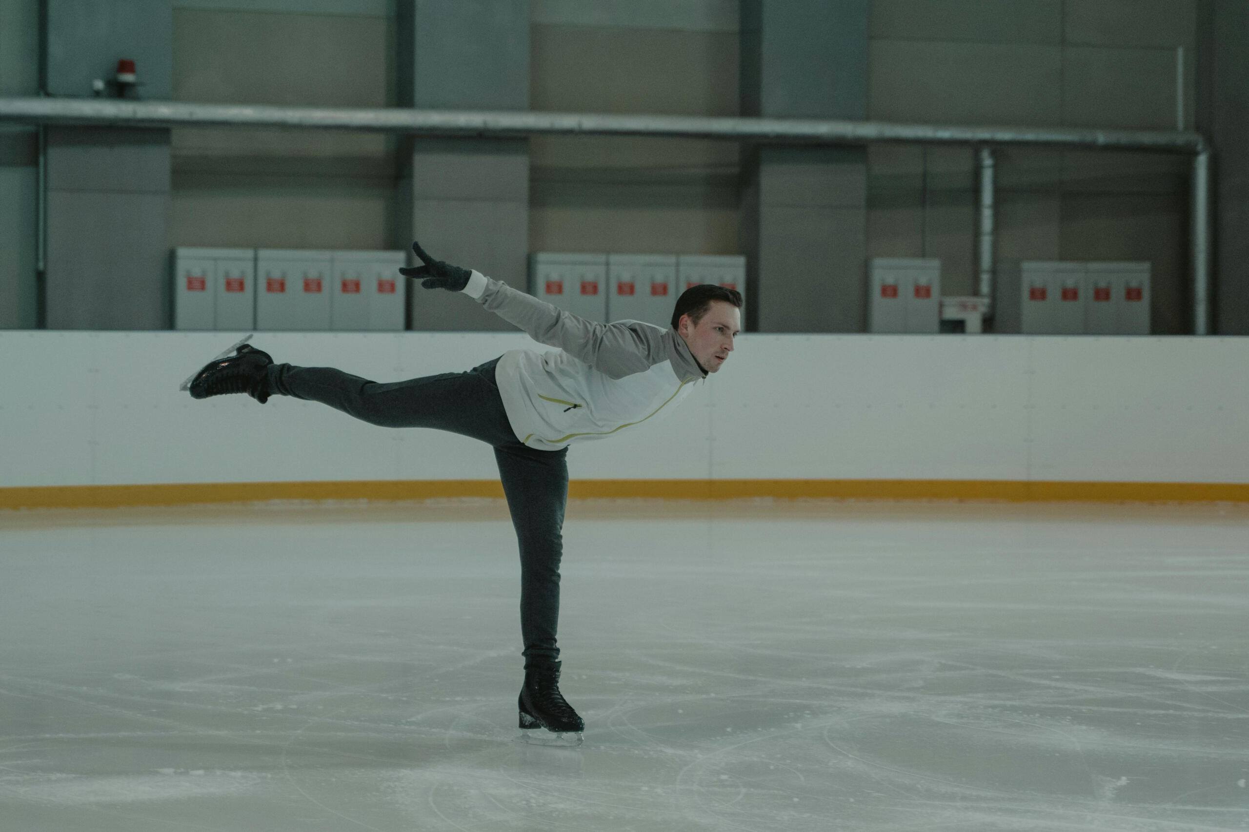 A man skates gracefully in an indoor rink. One leg is raised in a 90 degree angle.
