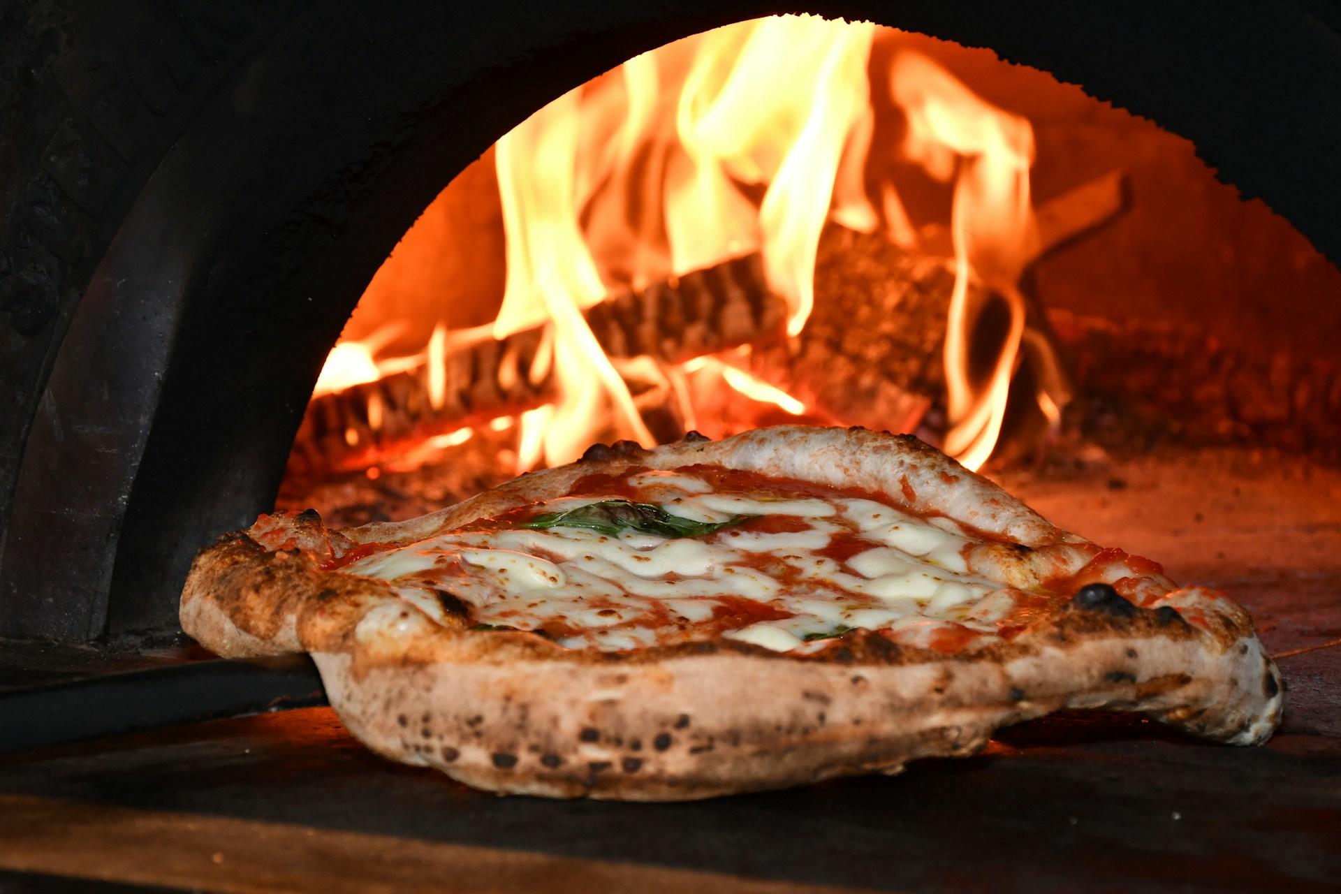 A pizza cooking inside a wood-fired oven with visible flames.