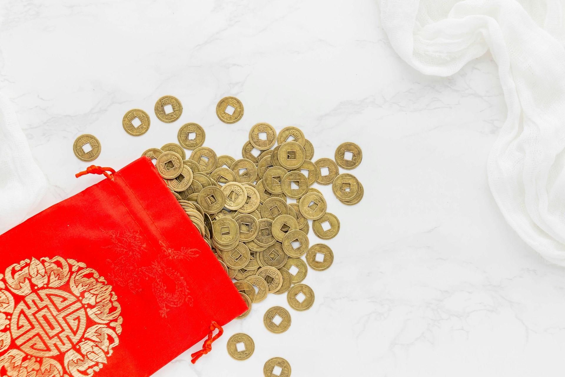 Chinese coins spilling from a red pouch on a marble surface with a white cloth.