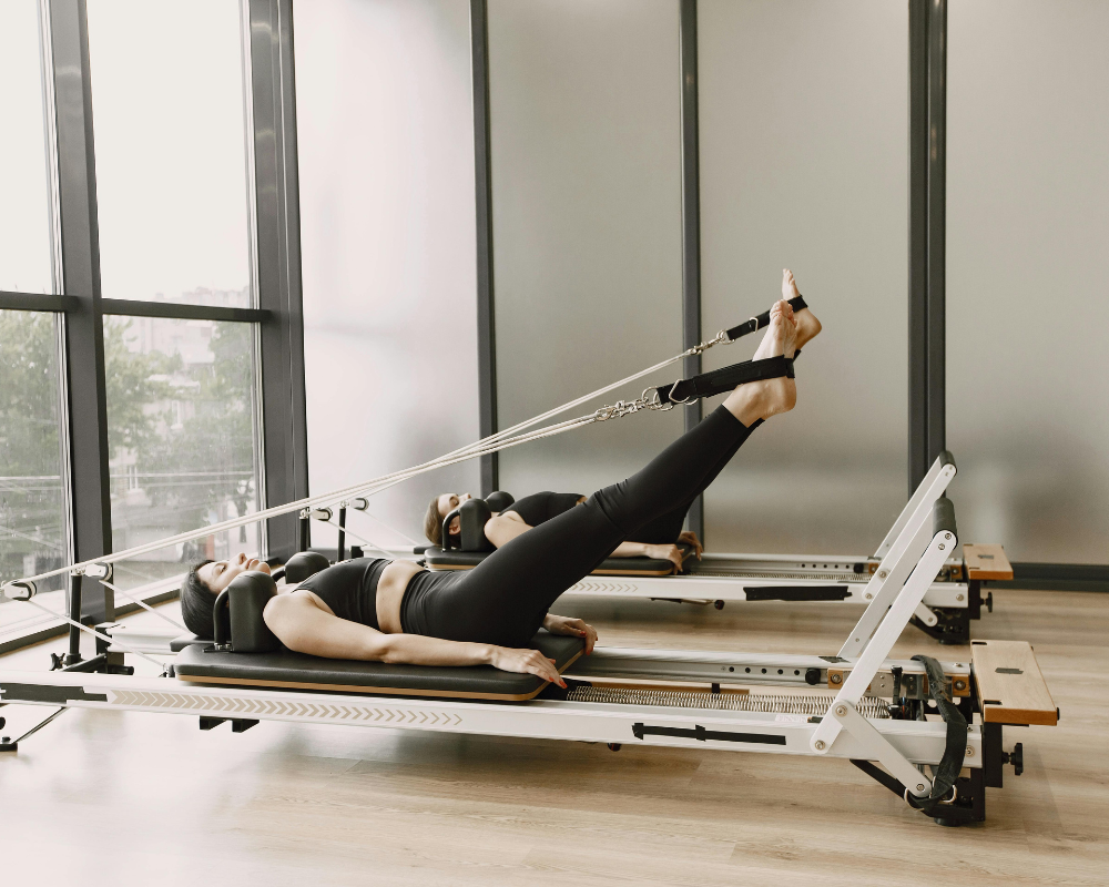 Two women performing leg stretches on reformer Pilates machines in a studio.