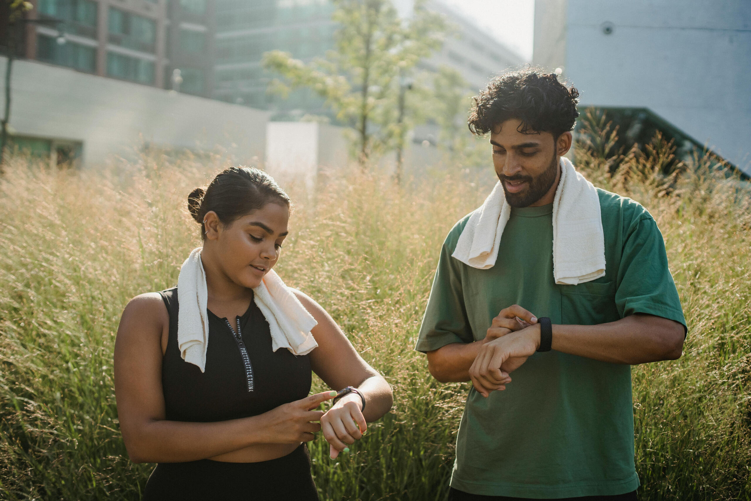 A man and woman look at their fitness watches. They stand in front of buildings. 