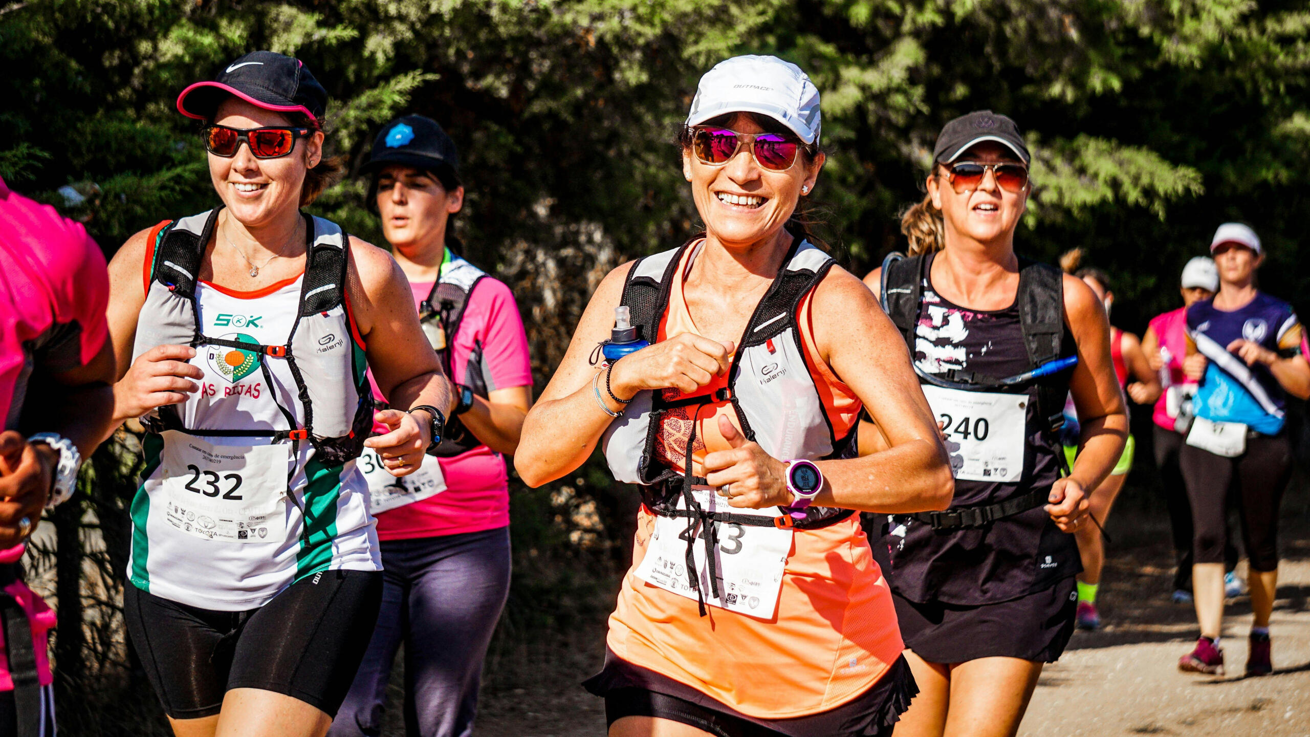 A group of women run in a marathon. They wear numbers and fitness gear.