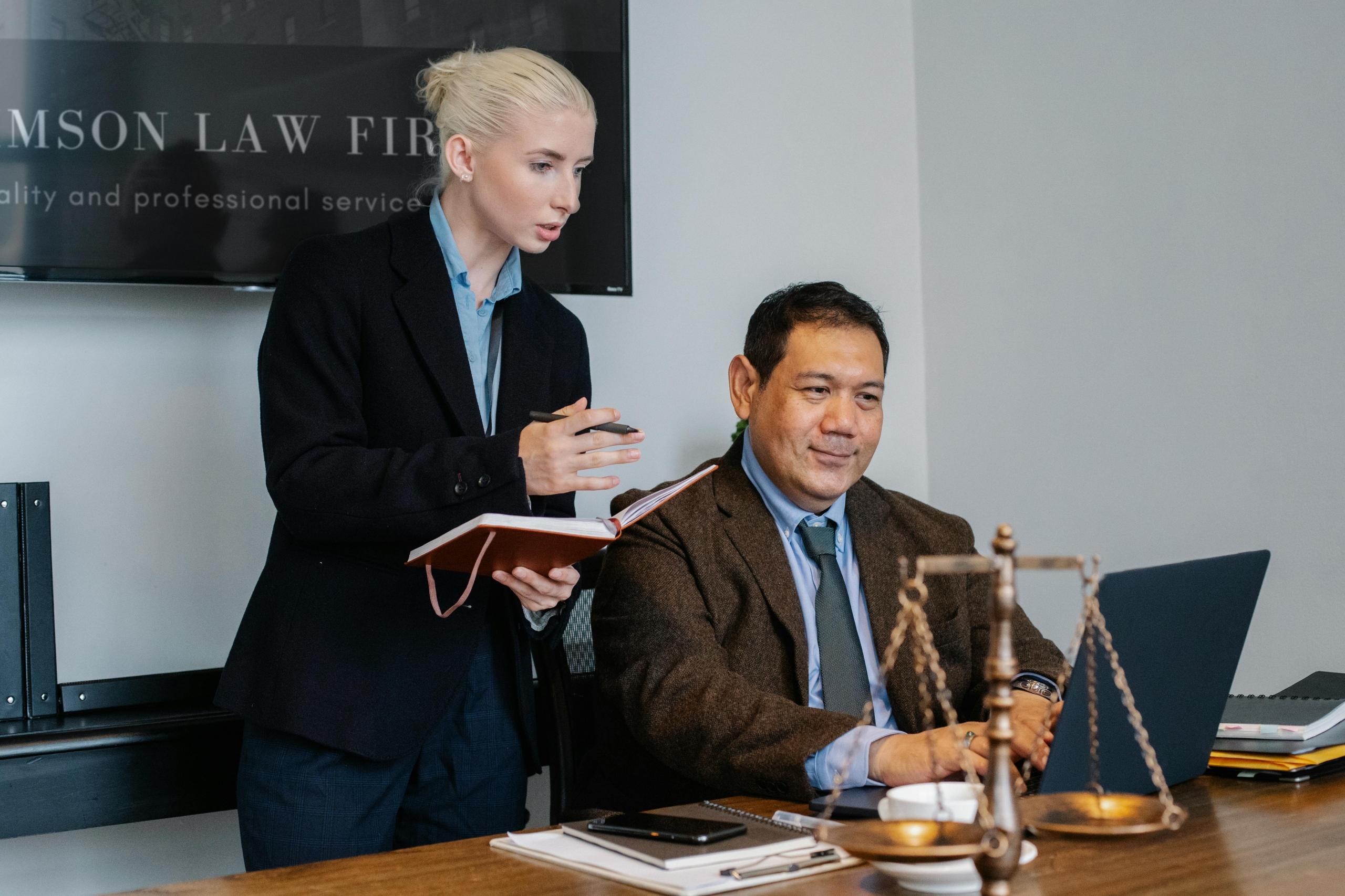 a woman stands beside a man talking notes in an office. The man looks at a laptop. The scales of justice can be seein on the desk.