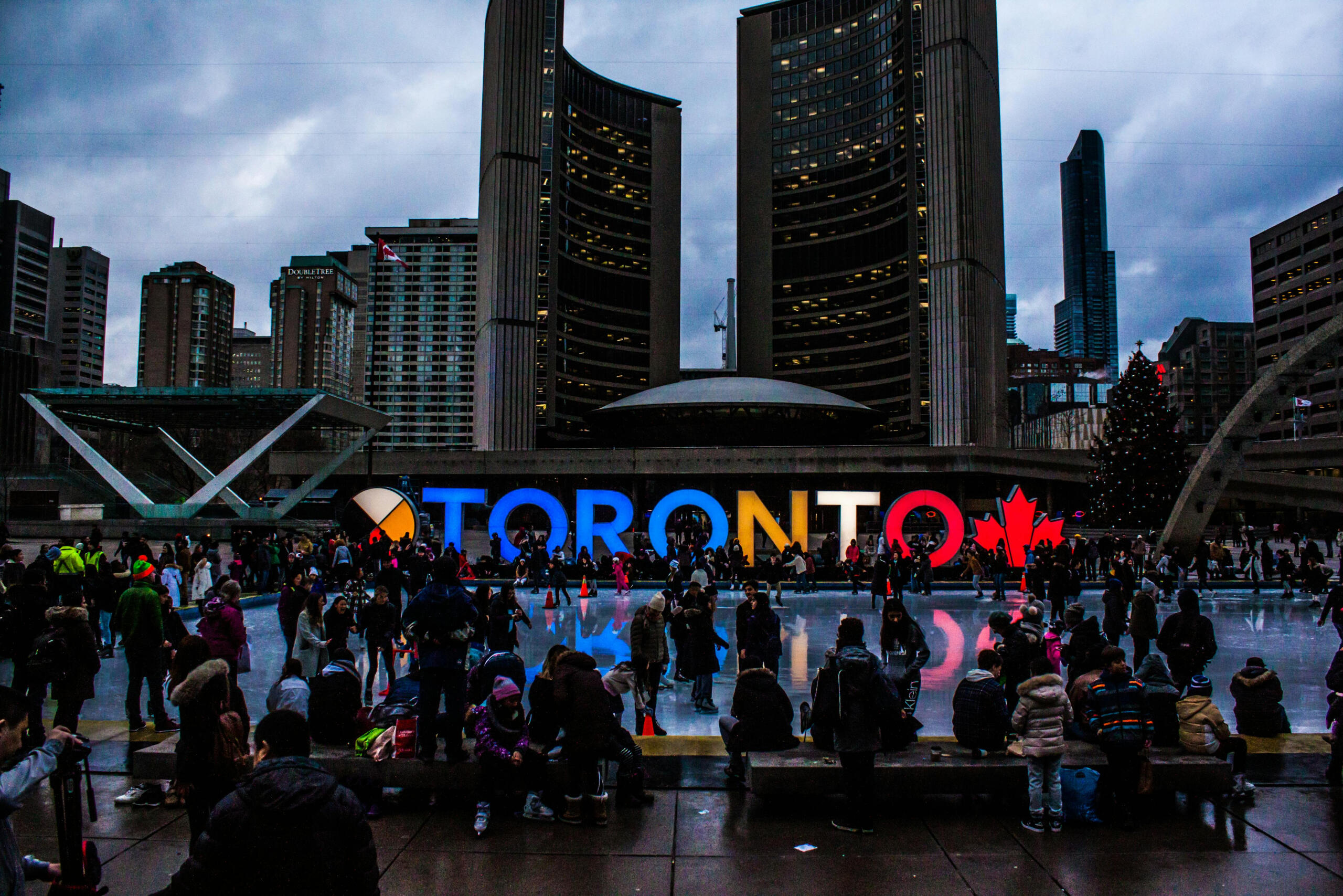 An ice skating rink filled with people in front of a sign that says Toronto. Two tall buildings are in the background.