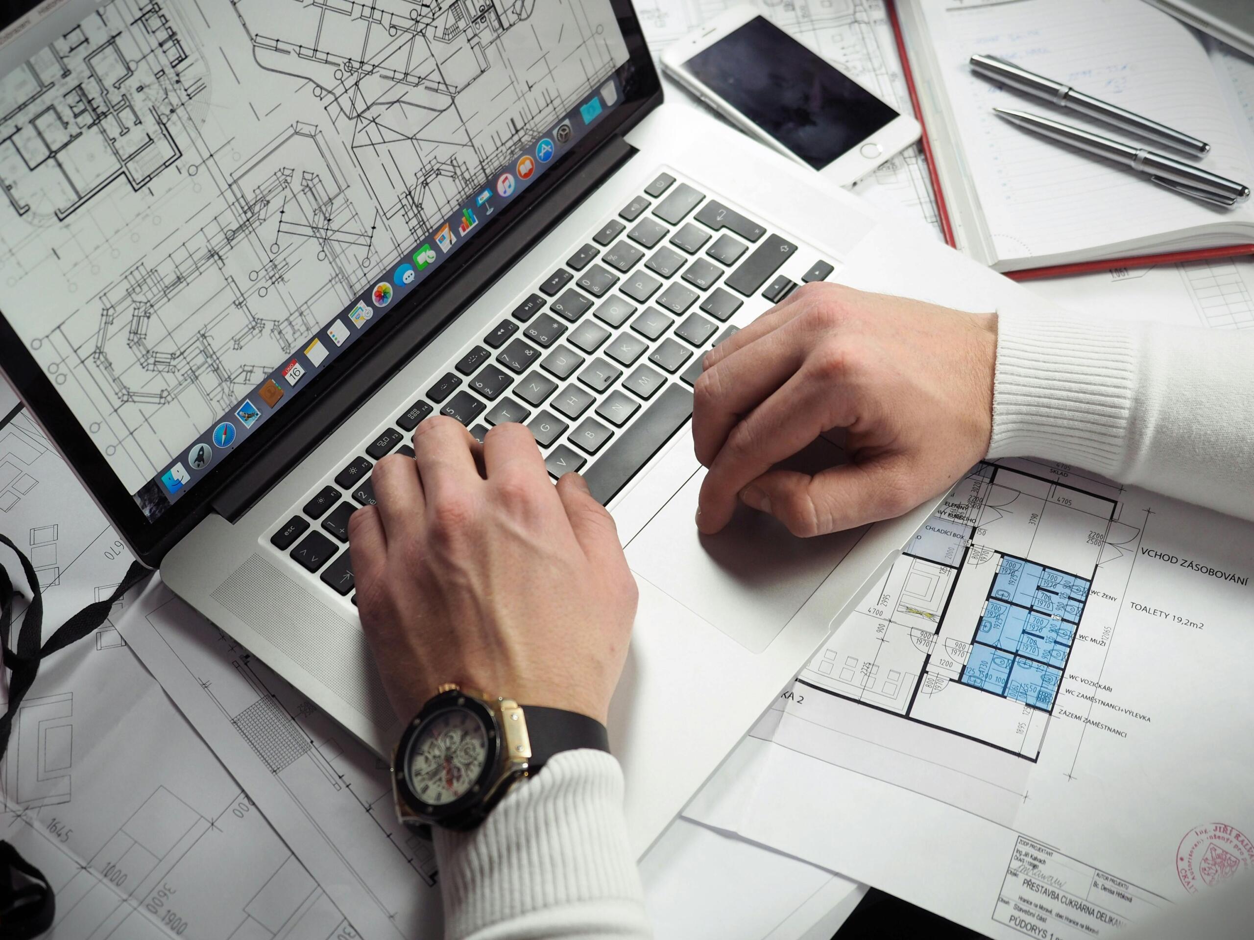A man works at a computer working on blueprints. Only his hands can be seen. Blueprints can be seen all over the desk.