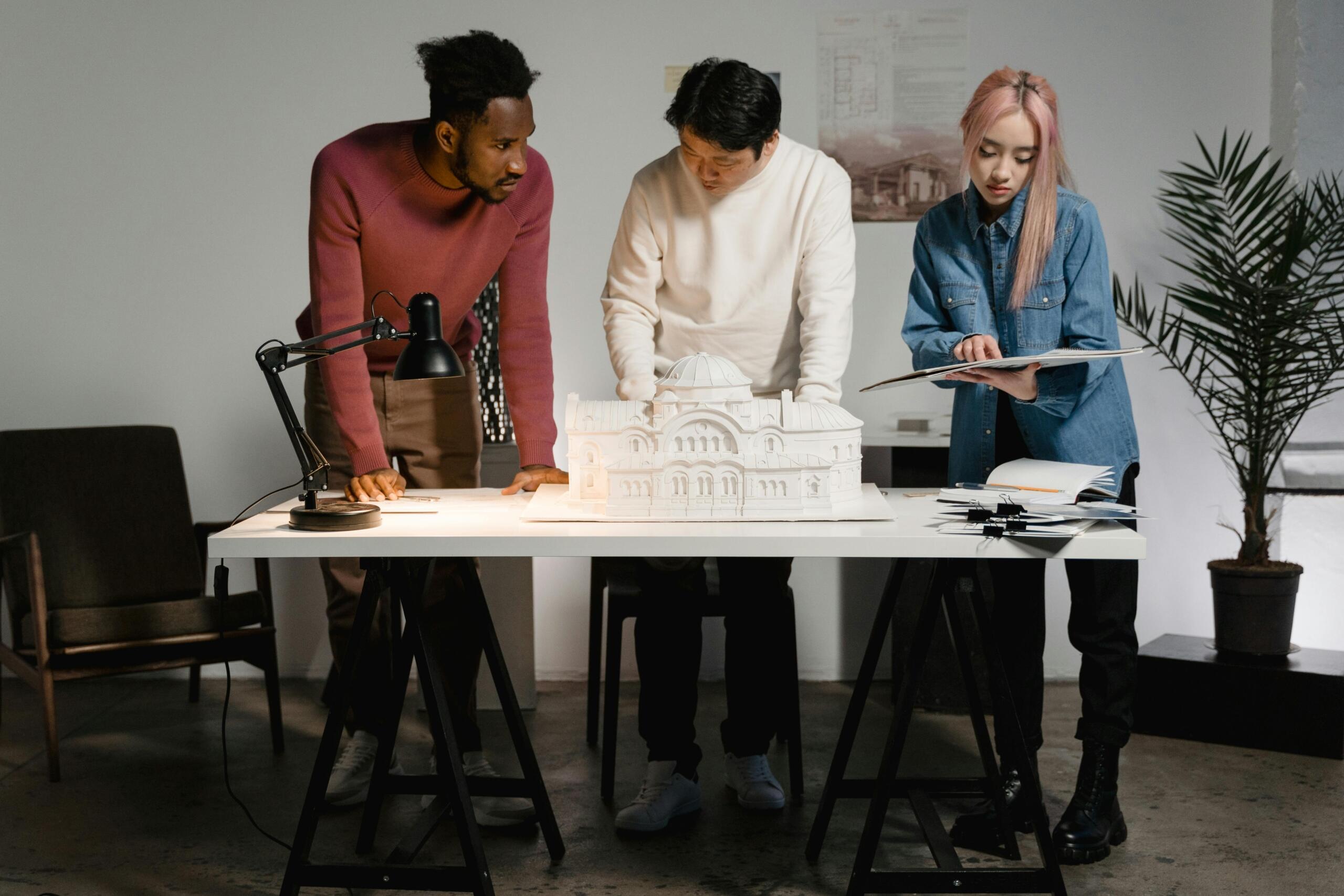 Three adult architecure students stand at a desk, looking at papers. A model of a building with a domed ceiling is on the desk.